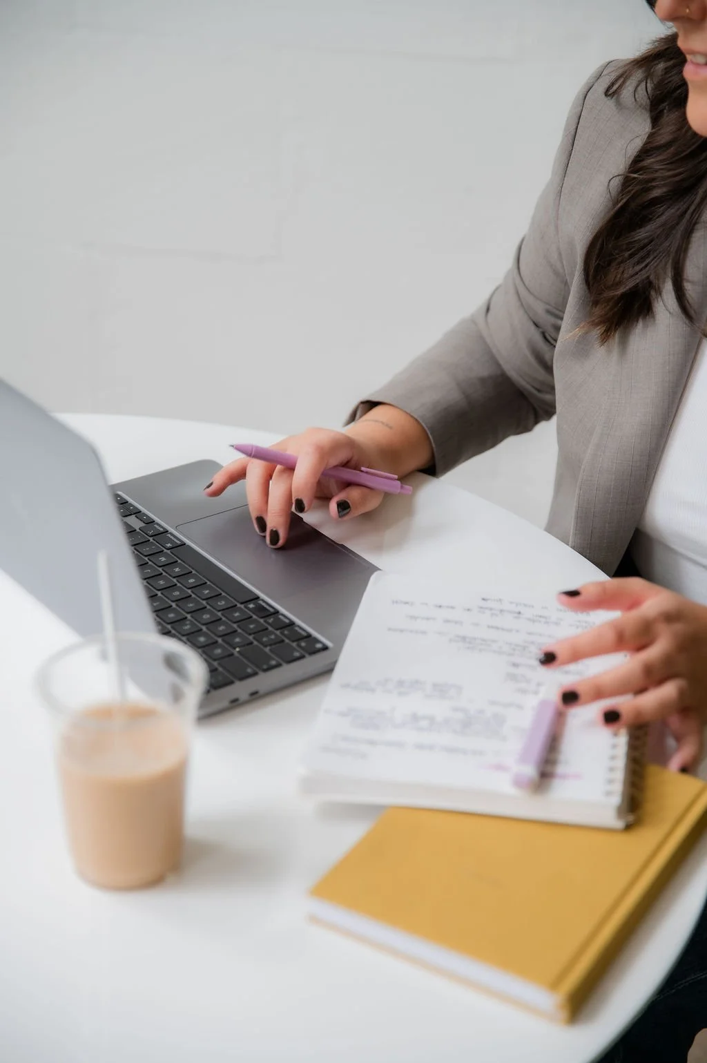 Person working at a white desk with a laptop, notes, a yellow notebook, and a drink.