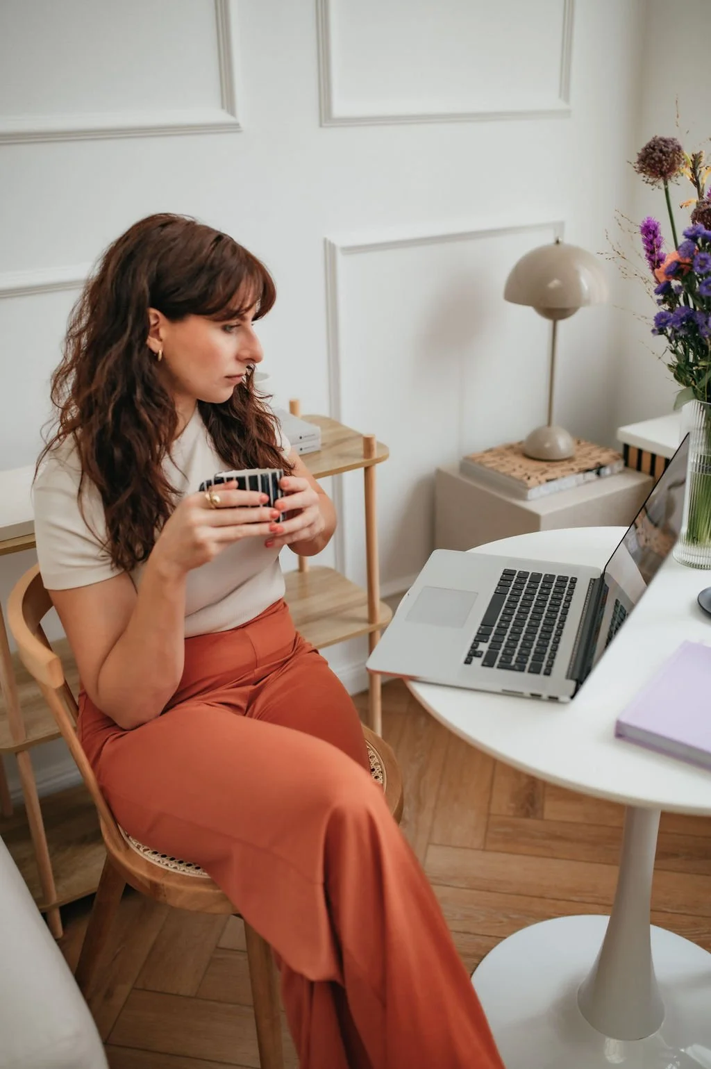 A woman sitting on a wooden chair holding a patterned mug, working on a laptop at a white table with a pink notebook. There is a side table with a lamp and flowers in the background.