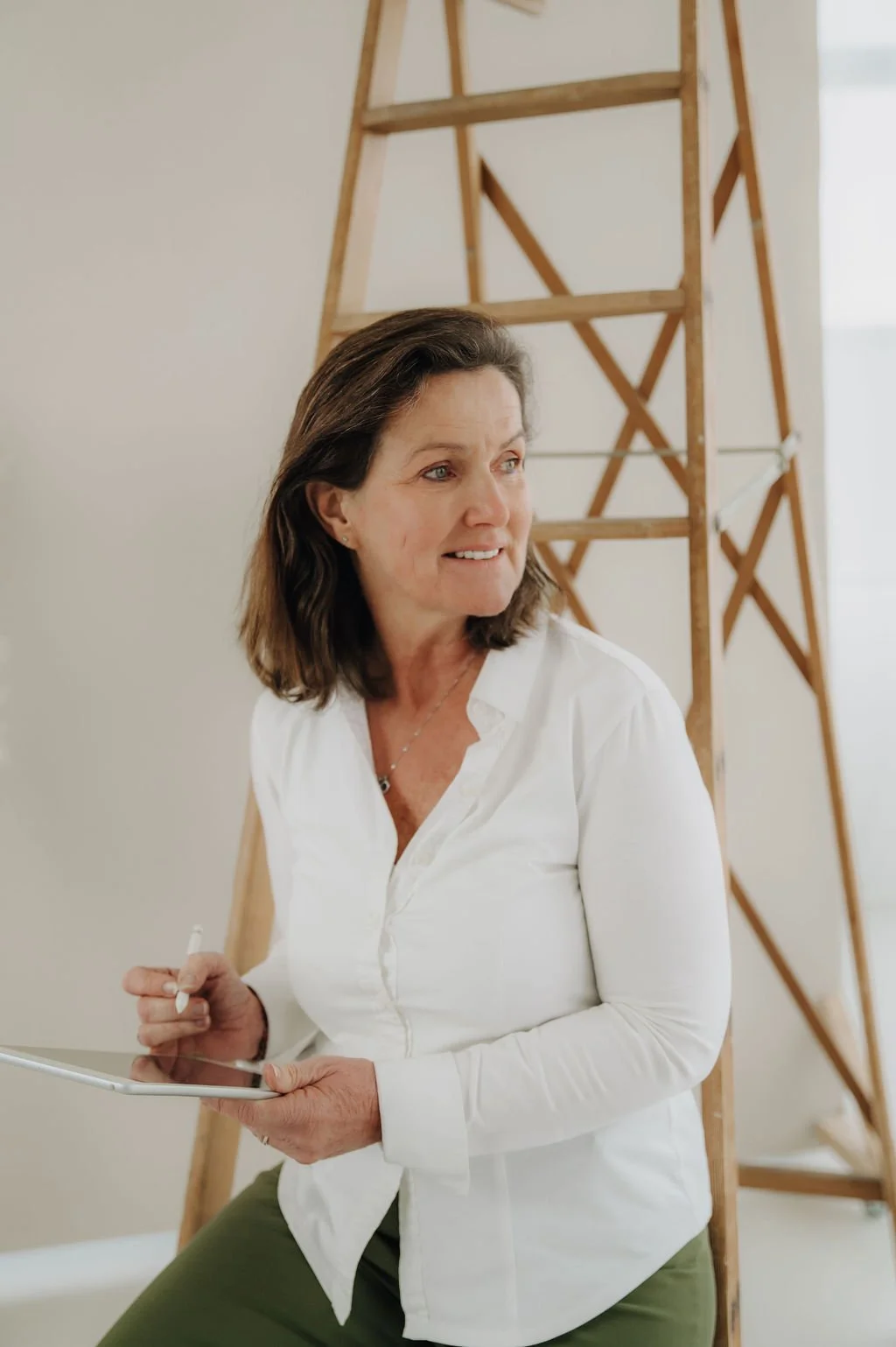 A woman with shoulder-length brown hair, wearing a white shirt and green pants, is sitting near a wooden ladder, holding a tablet and stylus, looking to her right.