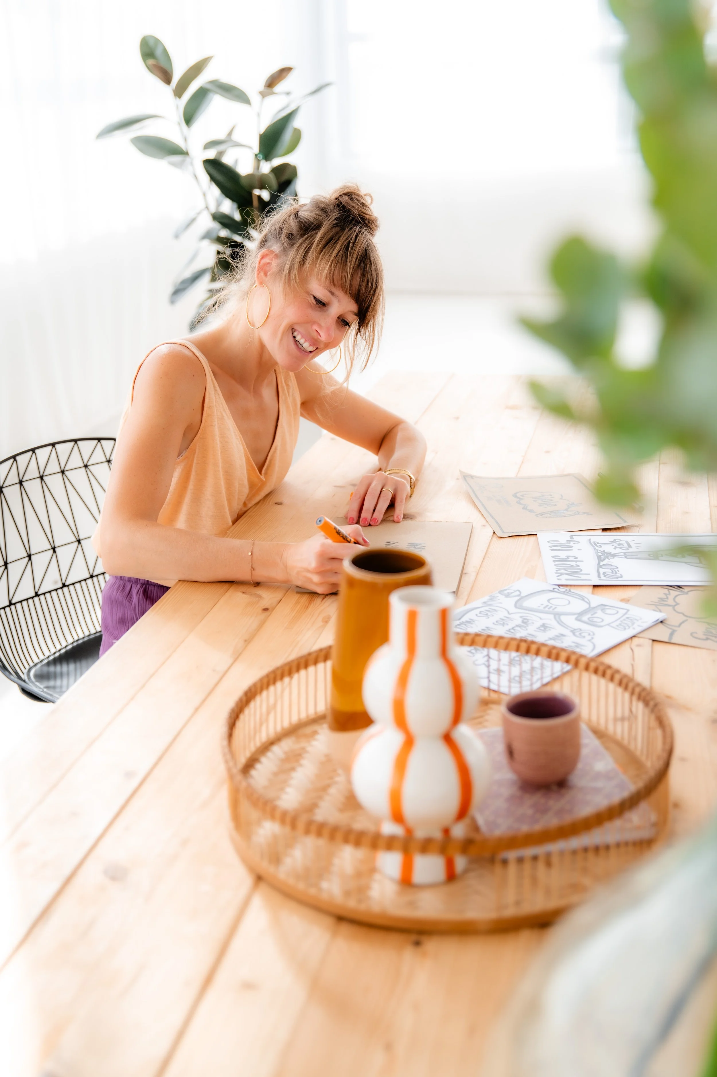 A woman sitting at a wooden table, smiling and drawing on paper with other drawings and notebooks around her. There are decorative pottery and vases on the table, and green plants in the background.