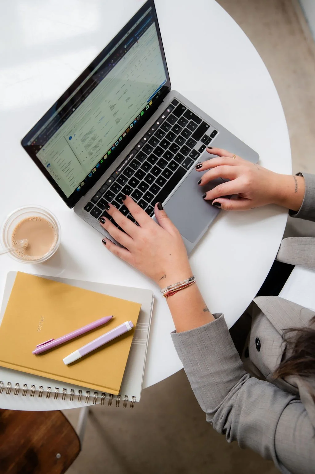 A person in a gray blazer using a silver MacBook laptop on a white round table, with a yellow notebook, pink and white pens, a spiral notebook, and a cup of coffee nearby.