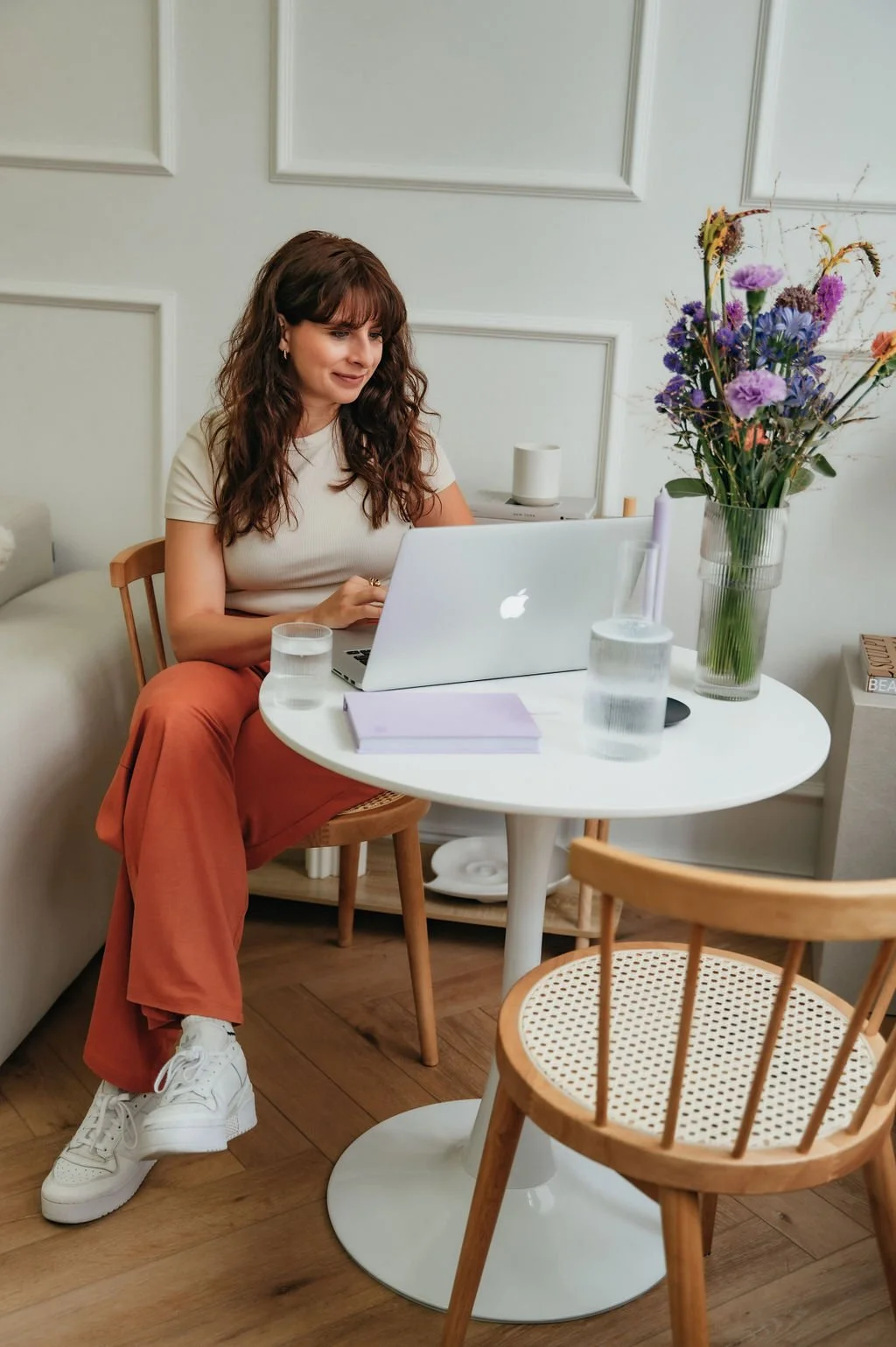 A woman with wavy brown hair, wearing a beige top and orange pants, sitting at a white table with a silver laptop, two glasses of water, a purple notebook, and a vase of purple and pink flowers inside a room with white walls and wooden floors.