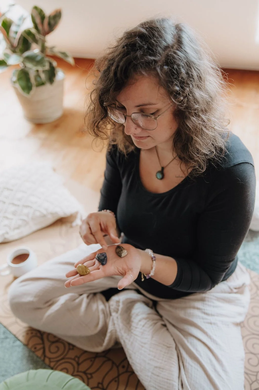 Woman sitting cross-legged on a rug, examining and holding four polished stones or crystals in her hand, with a potted plant and cup of coffee nearby.