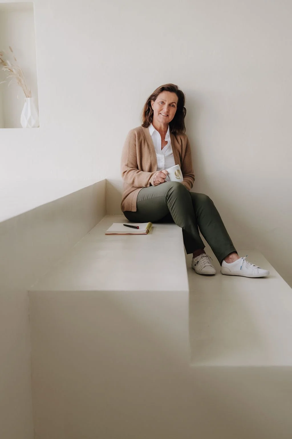 A woman sitting on a raised platform in a minimalist room, holding a mug, with a notebook and pen nearby.