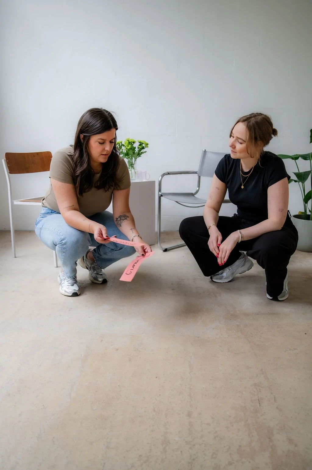 Two women squatting in a room with white walls, one holding a pink paper with handwritten words, the other observing. There are chairs and a potted plant in the background.