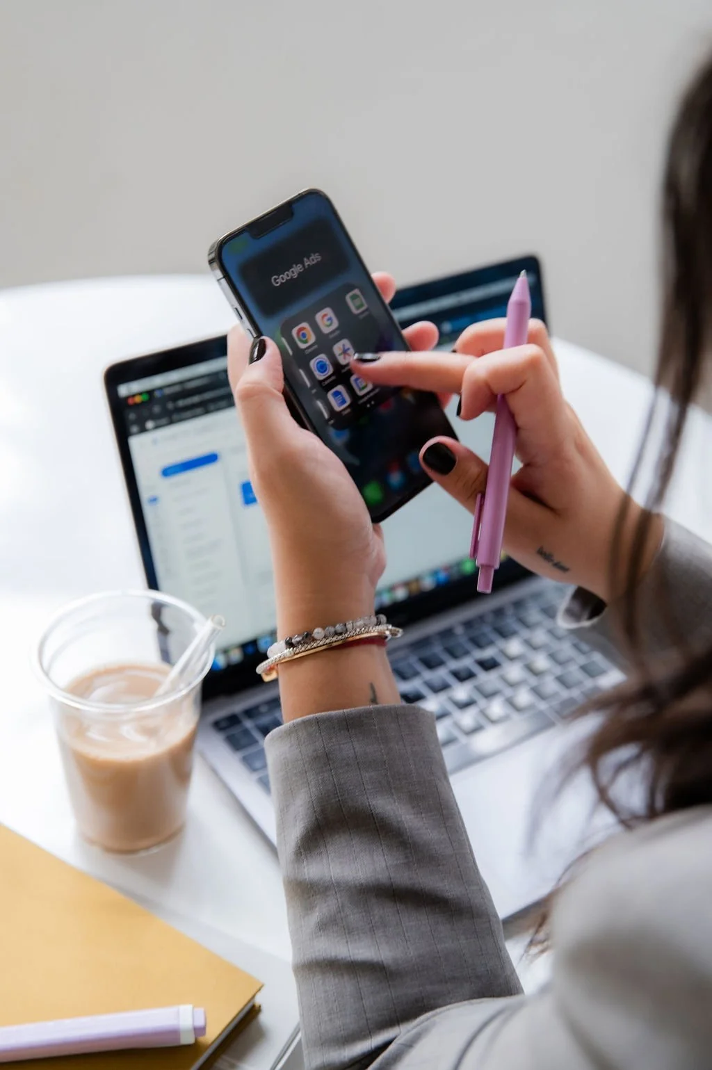 A person in a gray blazer holds a smartphone with a pink stylus, while working on a laptop with a document open. There is a cup of iced coffee, a yellow notebook, and a pink pen on the white desk.