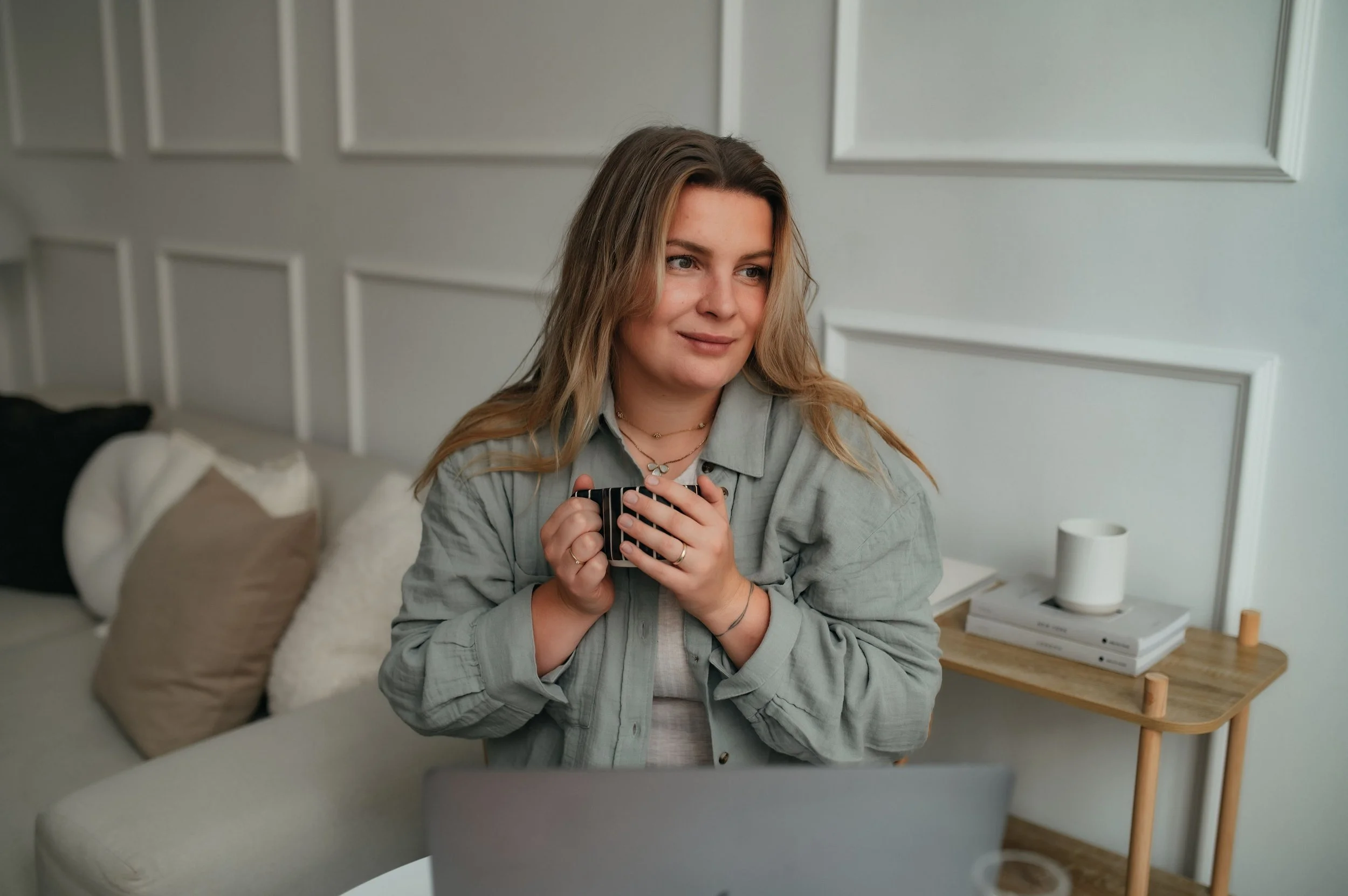 A woman with long wavy hair holding a mug, sitting in front of a laptop on a table, with a white wall and side table with books, a lamp, and a cup in the background.