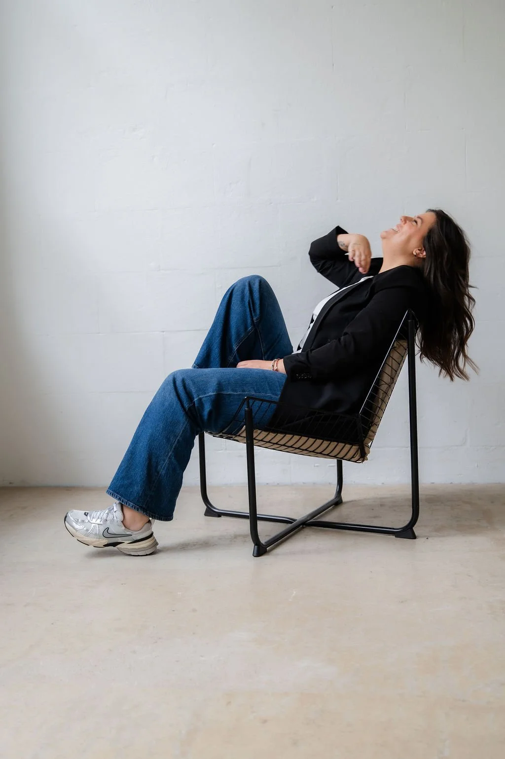 Woman sitting on a wire-framed chair, leaning back and laughing, in a room with plain white walls and light-colored floor.