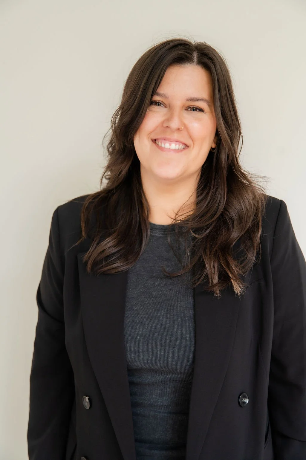 A woman with shoulder-length brown hair, smiling, wearing a black blazer over a gray top, standing against a plain light-colored background.
