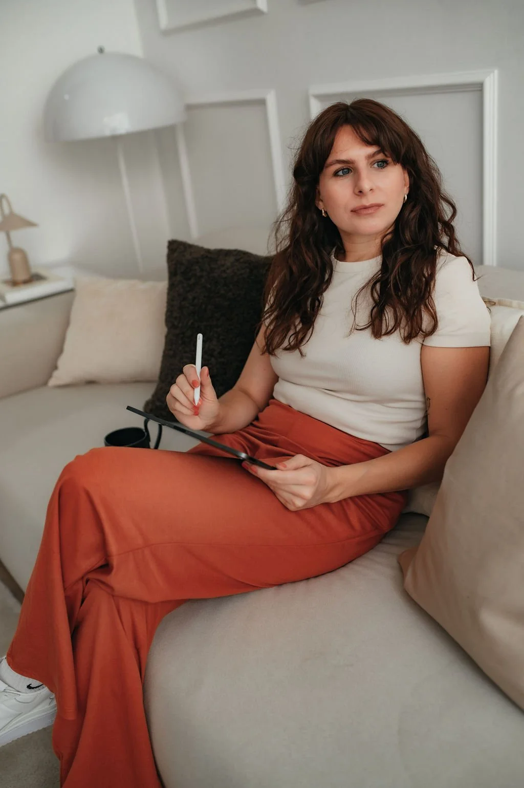 A woman sitting on a light-colored sofa, holding a stylus and a digital drawing tablet, with pillows and a lamp on a side table in the background.