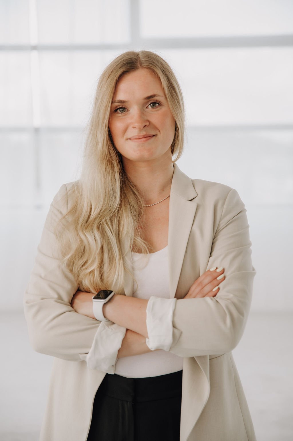 A smiling woman with long blonde hair wearing a beige blazer, white top, and a watch, standing with her arms crossed in front of a bright, modern background.