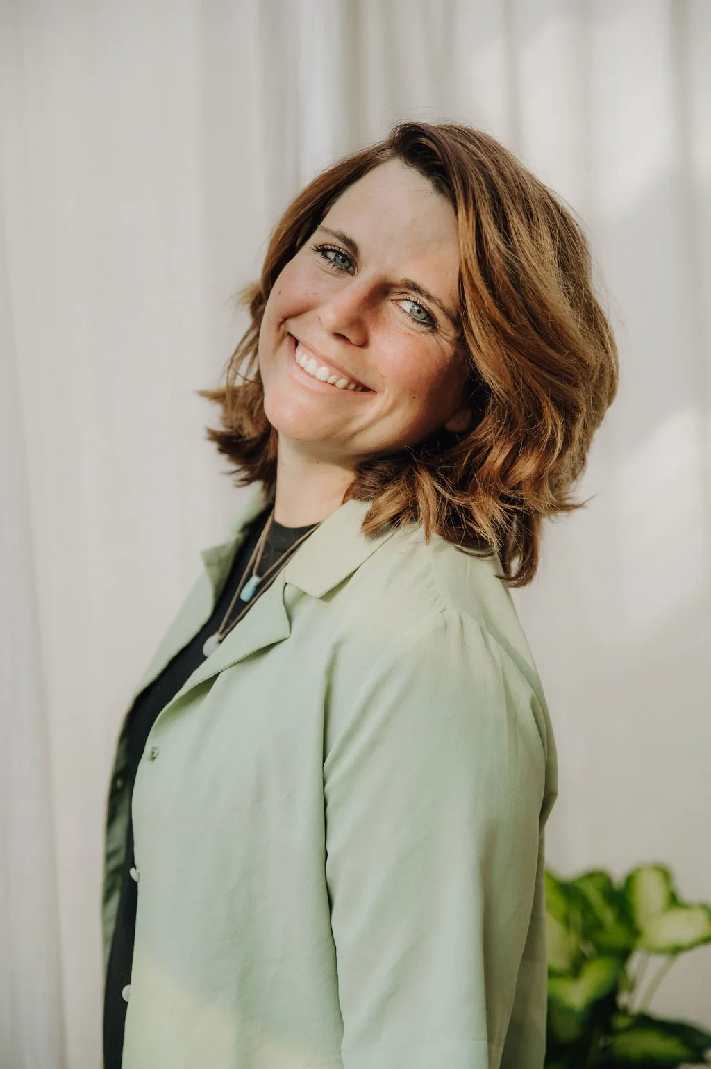 A woman with shoulder-length reddish-brown hair smiling with teeth, wearing a light green jacket over a black top, standing indoors near a light-colored wall with a plant in the background.