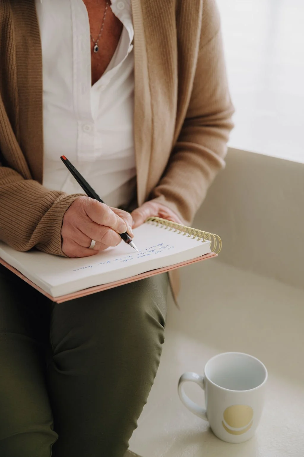 Person sitting on a couch, writing notes in a spiral notebook with a black pen, wearing a beige blazer and white shirt, with a white coffee mug nearby.