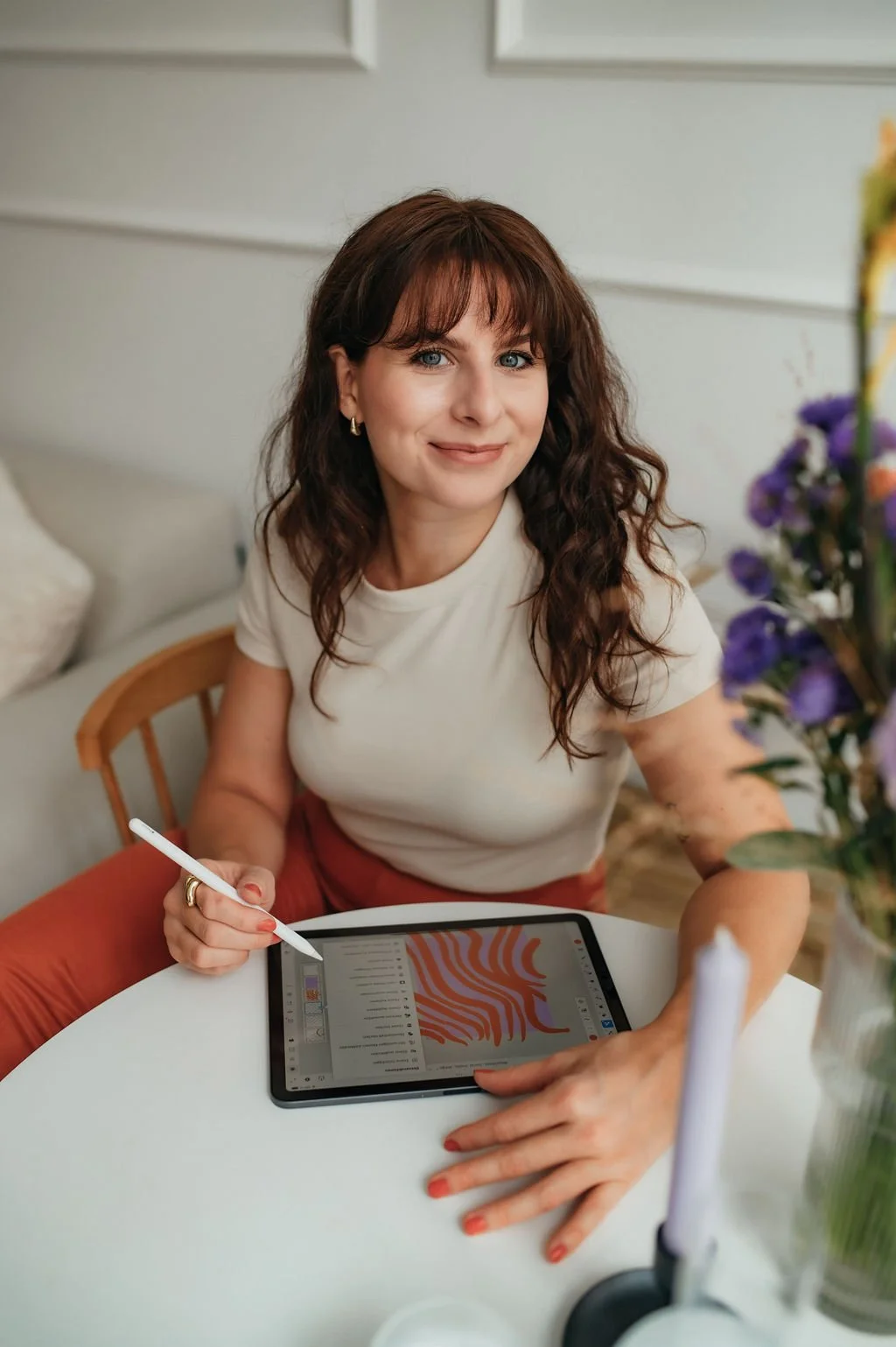 A woman with brown hair and blue eyes sitting at a white table with a tablet and stylus, smiling at the camera, with flowers in the foreground.