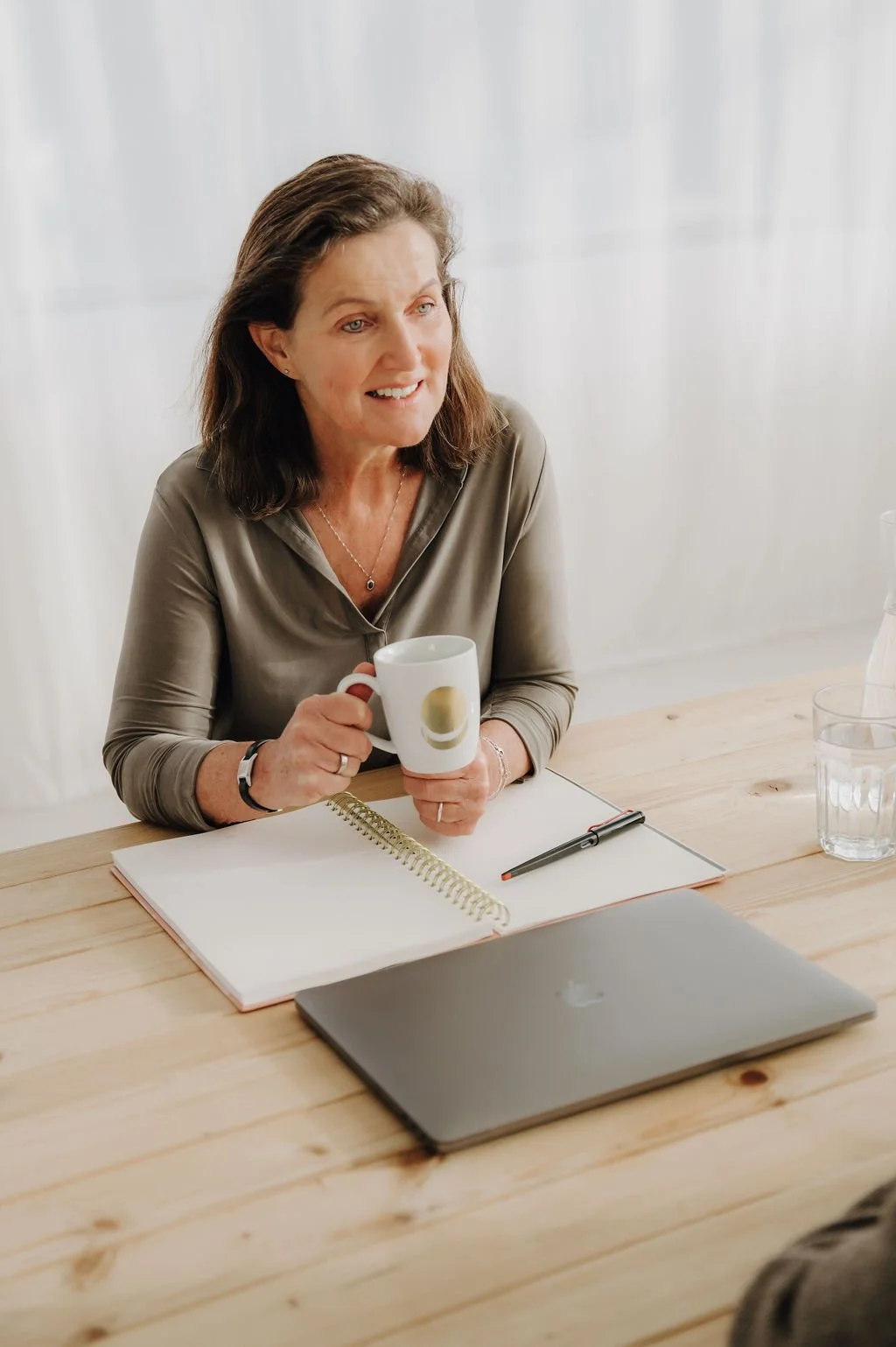 A woman sitting at a wooden table, holding a white coffee mug, with an open notebook, a pen, a closed laptop, and a glass of water in front of her, during a meeting or discussion.