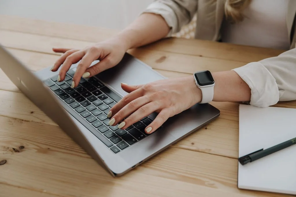 Person working on a laptop at a wooden table with a notebook and pen nearby, wearing a smartwatch.
