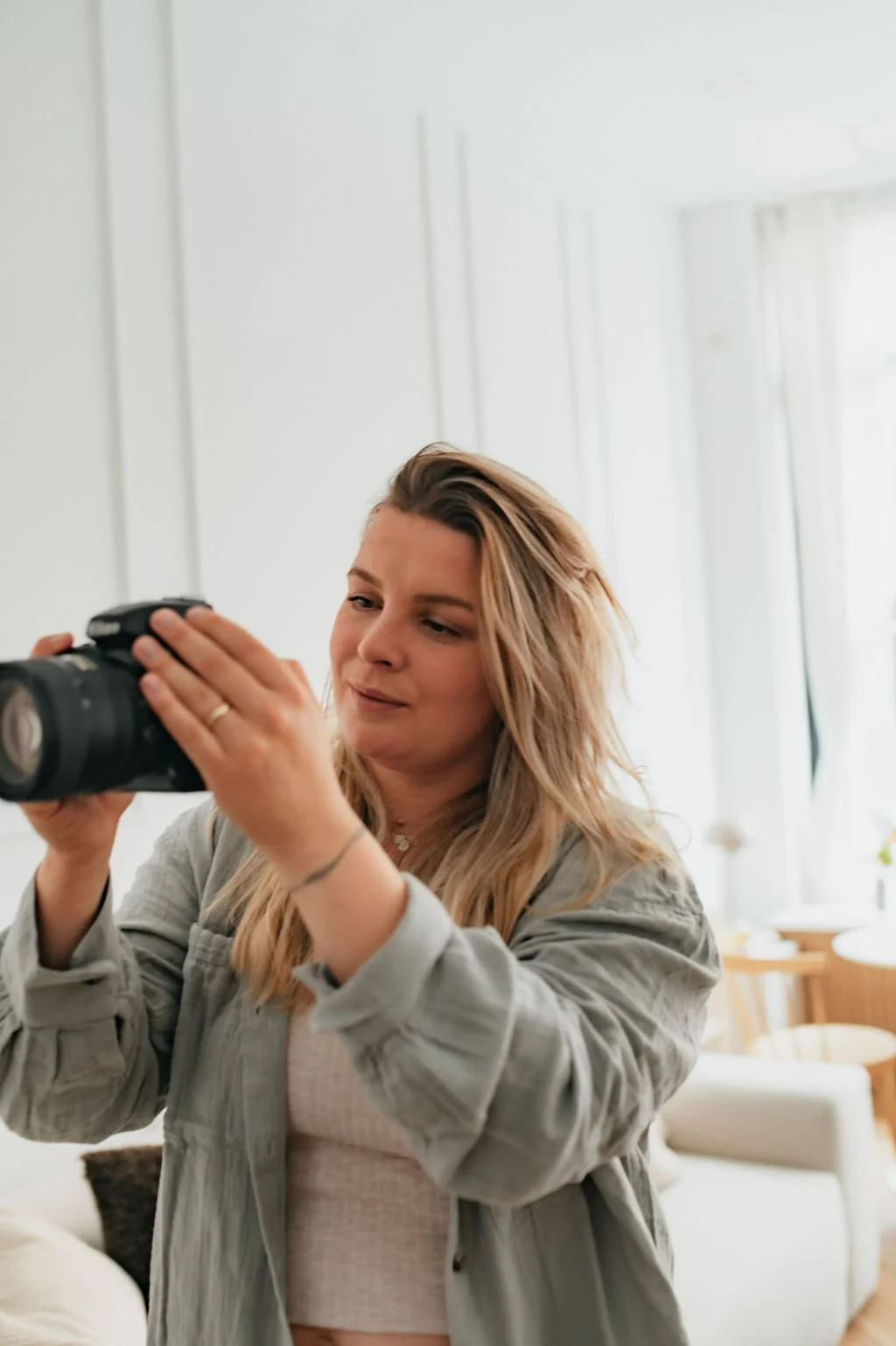A woman with wavy blonde hair is holding a camera and looking at it, inside a bright room with white walls and large windows.
