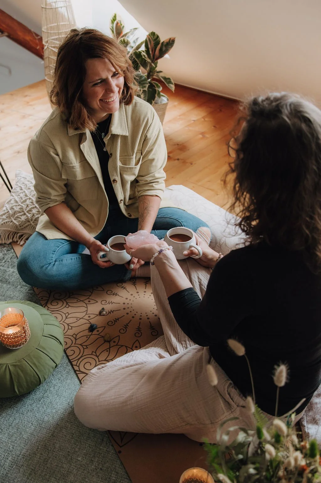 Two women sitting on the floor having a tea ceremony, exchanging mugs, with candles and plants in a cozy, decorated room.