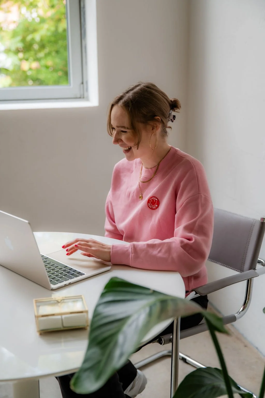 A young woman with short brown hair, wearing a pink sweatshirt, is sitting at a white desk and smiling while working on a laptop. There is a window behind her showing green trees outside, and a plant in the foreground.