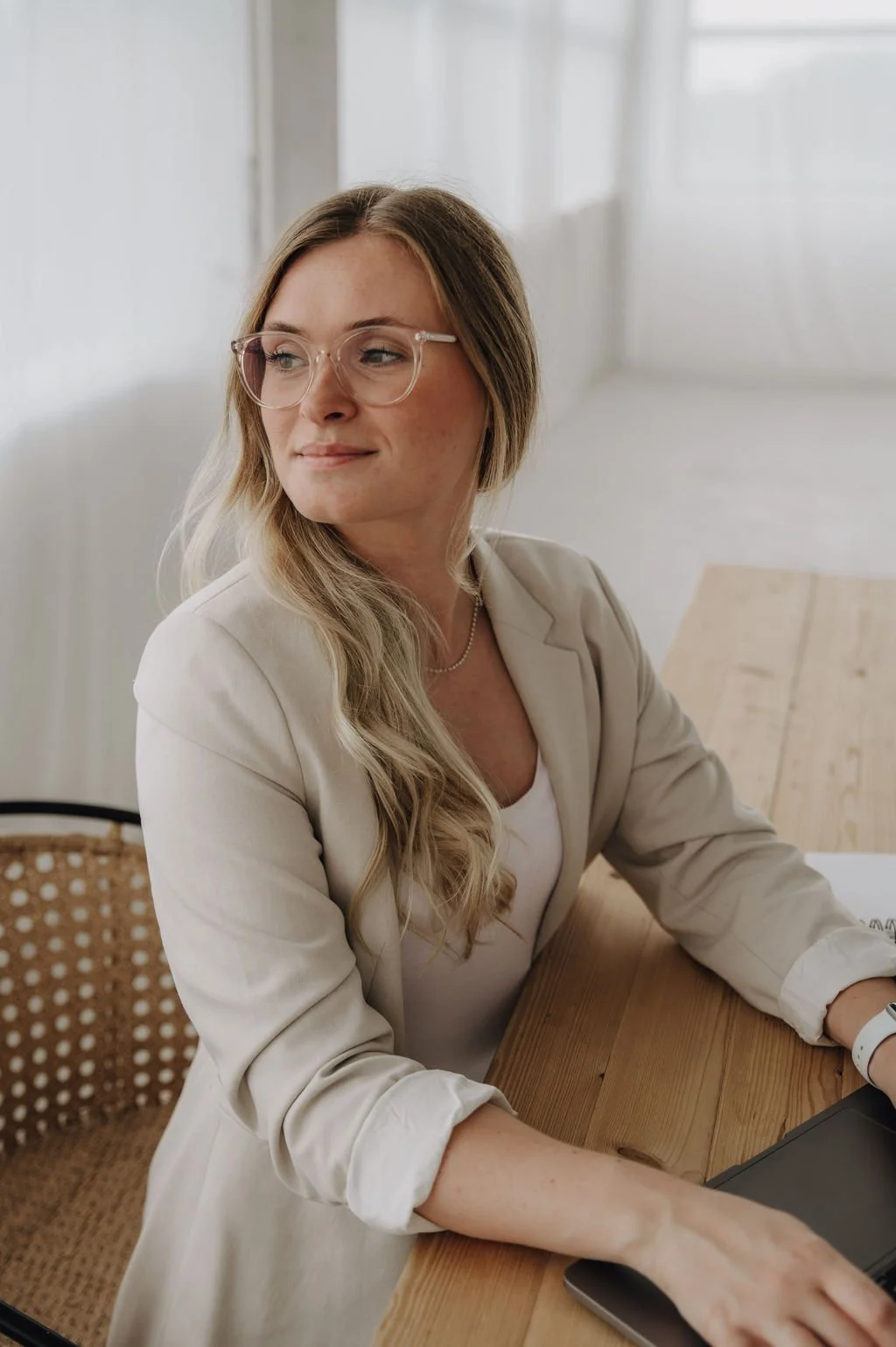 A woman with long blonde hair, glasses, and a beige blazer sitting at a wooden table with a laptop.