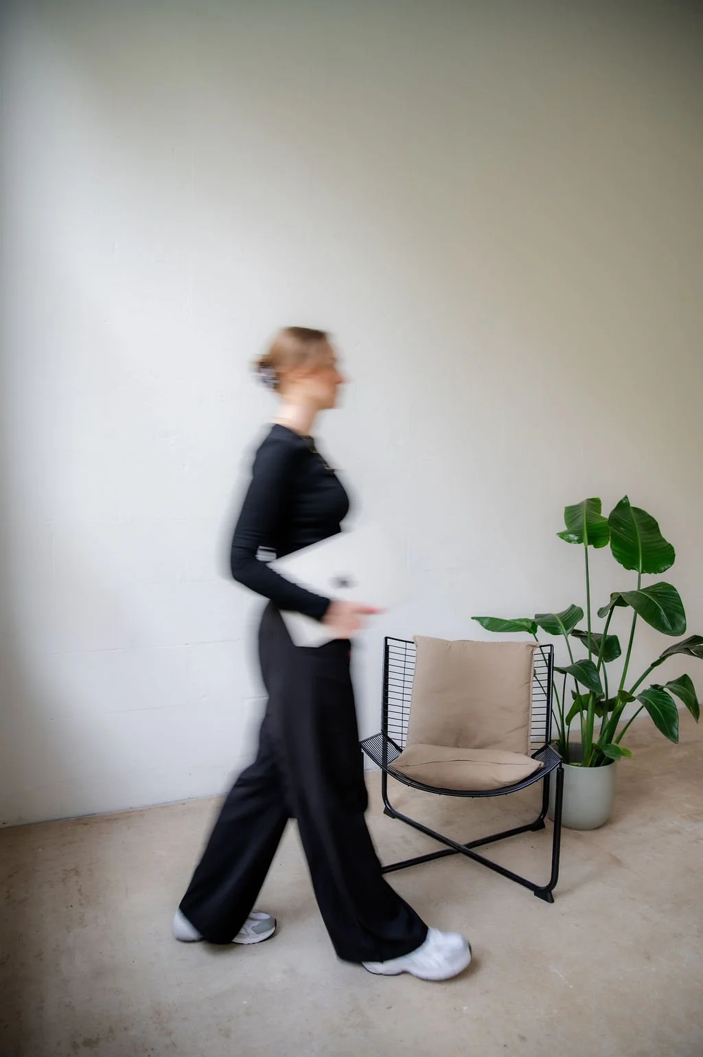 A woman walking past a beige chair and large green plant in a white pot against a plain wall.
