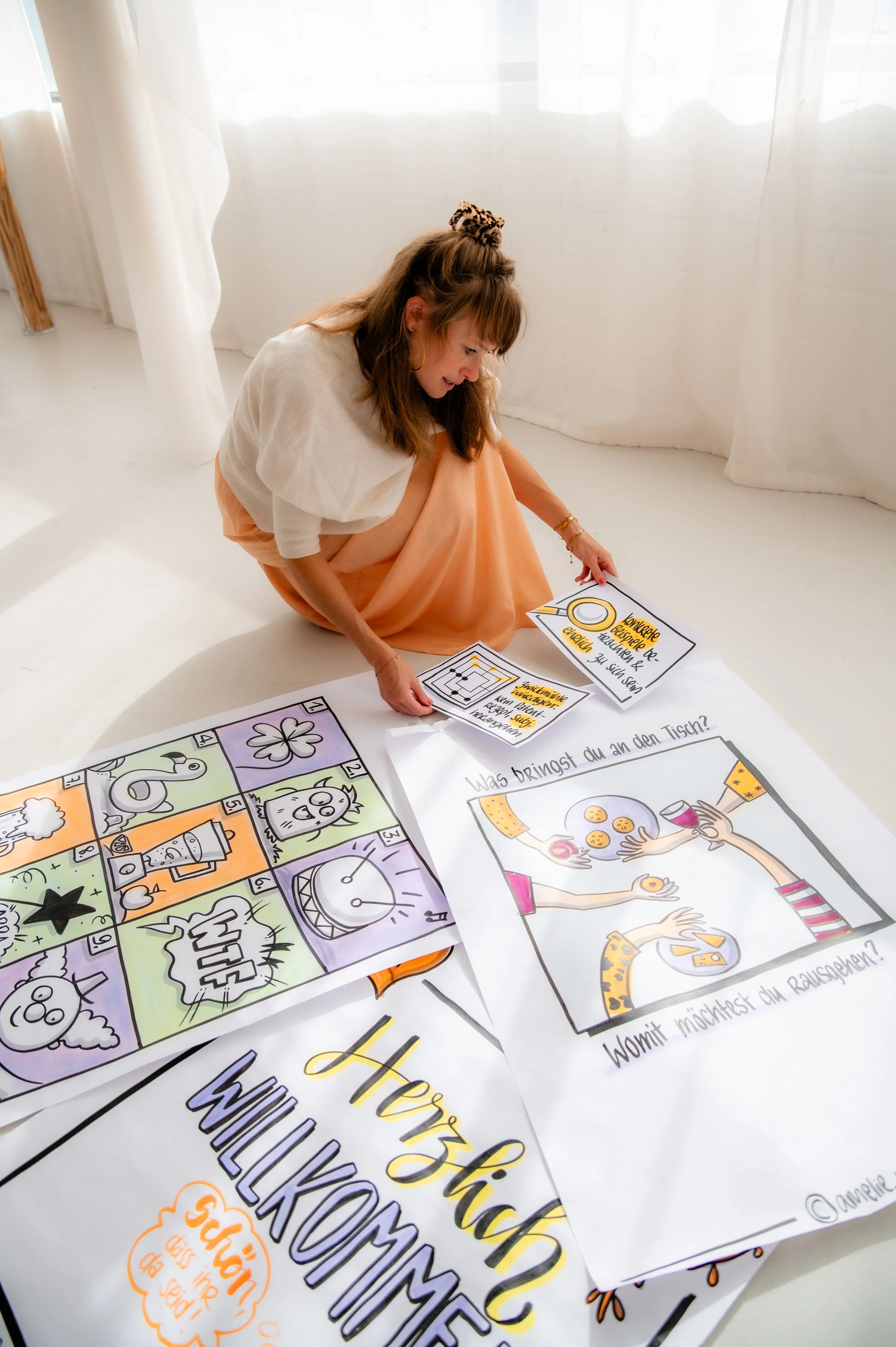 Woman with brown hair in a peach skirt and white top sitting on the floor, looking at colorful illustrated posters and papers with German text, in a bright room with white curtains.