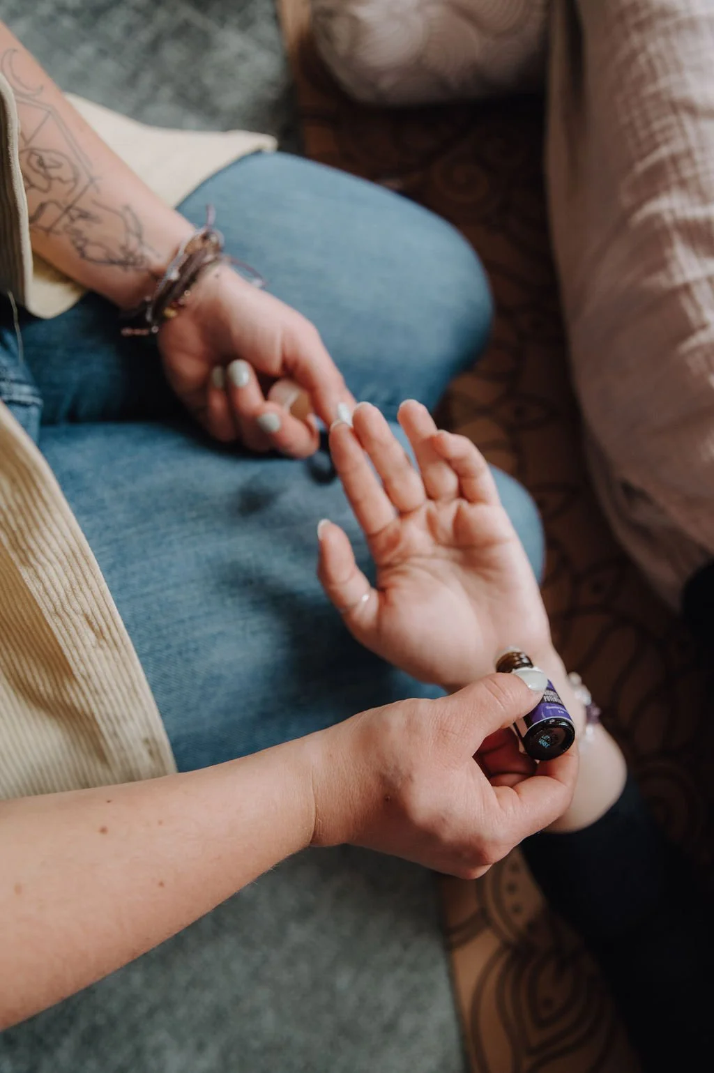 A person is applying essential oil or serum to another person's hand using a small brown bottle. The person receiving the treatment is sitting on a chair with blue jeans, and their hand is open, palm up. The setting appears to be casual, possibly at a wellness or self-care session.