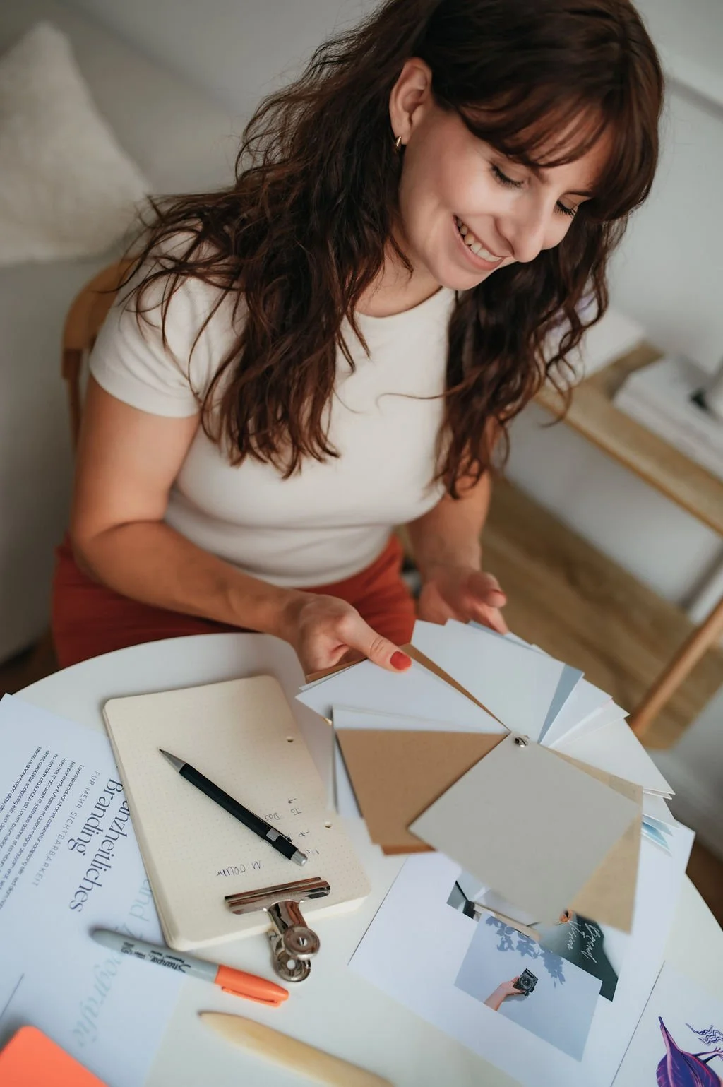 A woman smiling and looking down at color samples and design materials on a table, with a notebook, pen, and various papers.