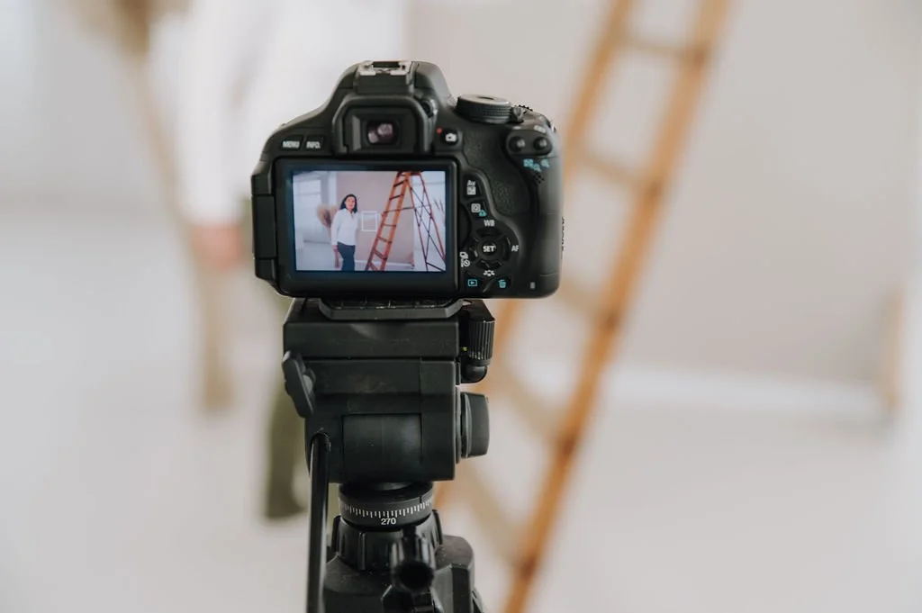 A DSLR camera on a tripod capturing an indoor photo of a woman standing next to a ladder.