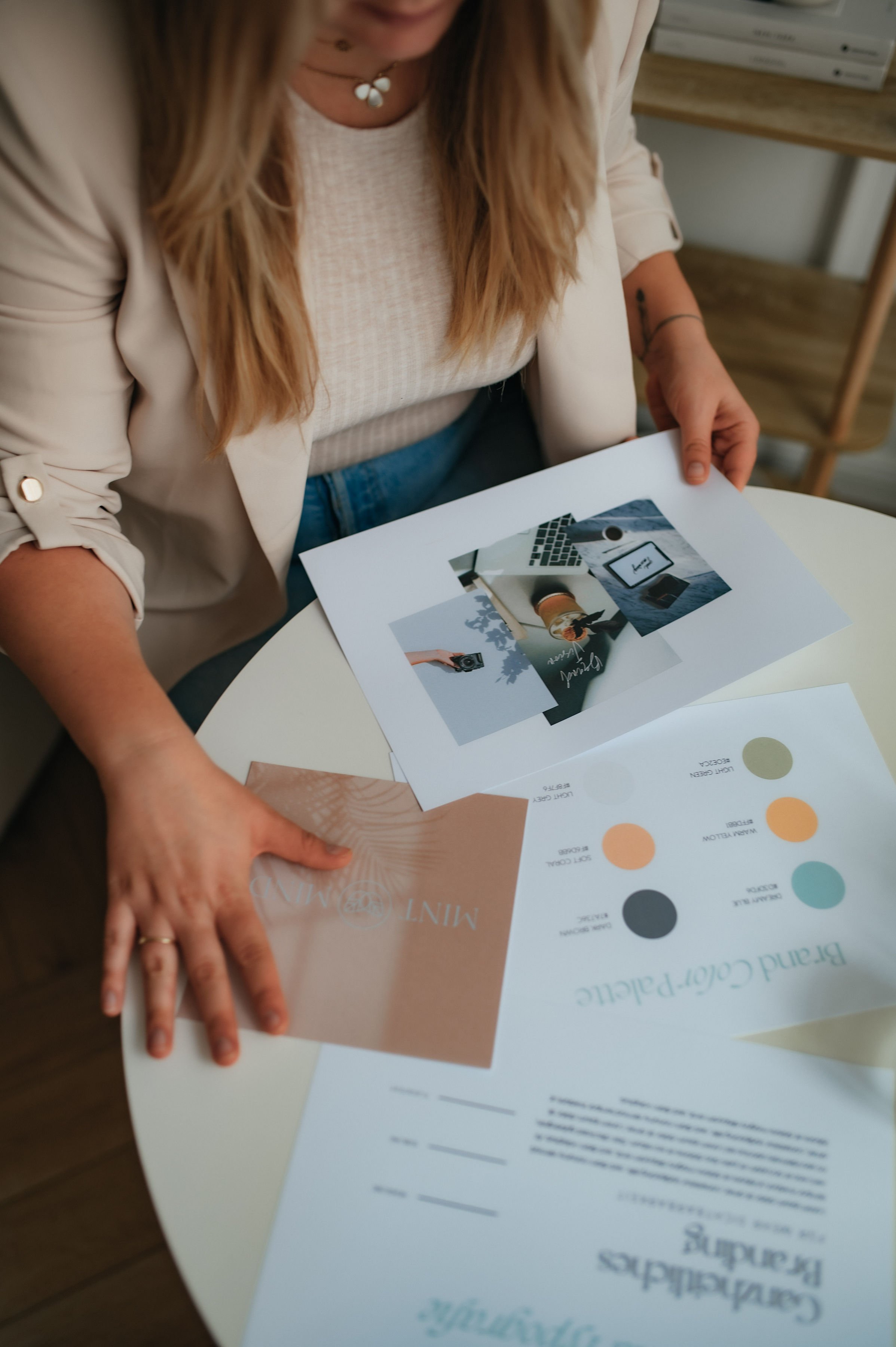 A woman sitting at a white table reviewing printed photos, color palette, and informational sheet for a branding project.