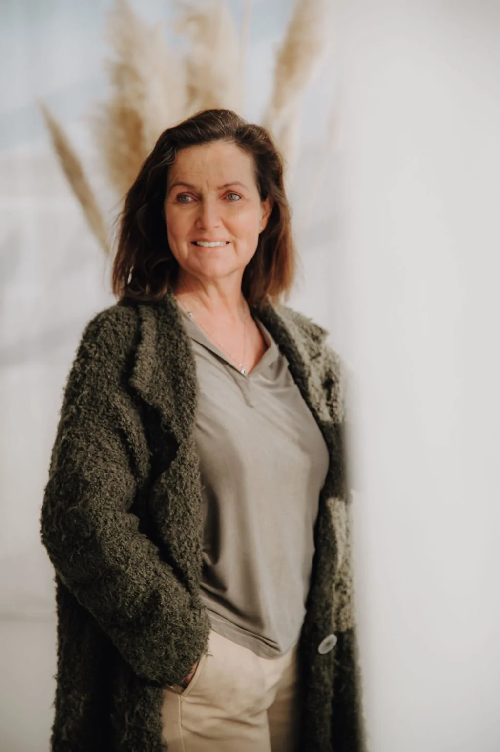 A woman with brown hair wearing a beige shirt, beige pants, and a dark, fluffy jacket, standing in front of a light-colored wall with decorative pampas grass.