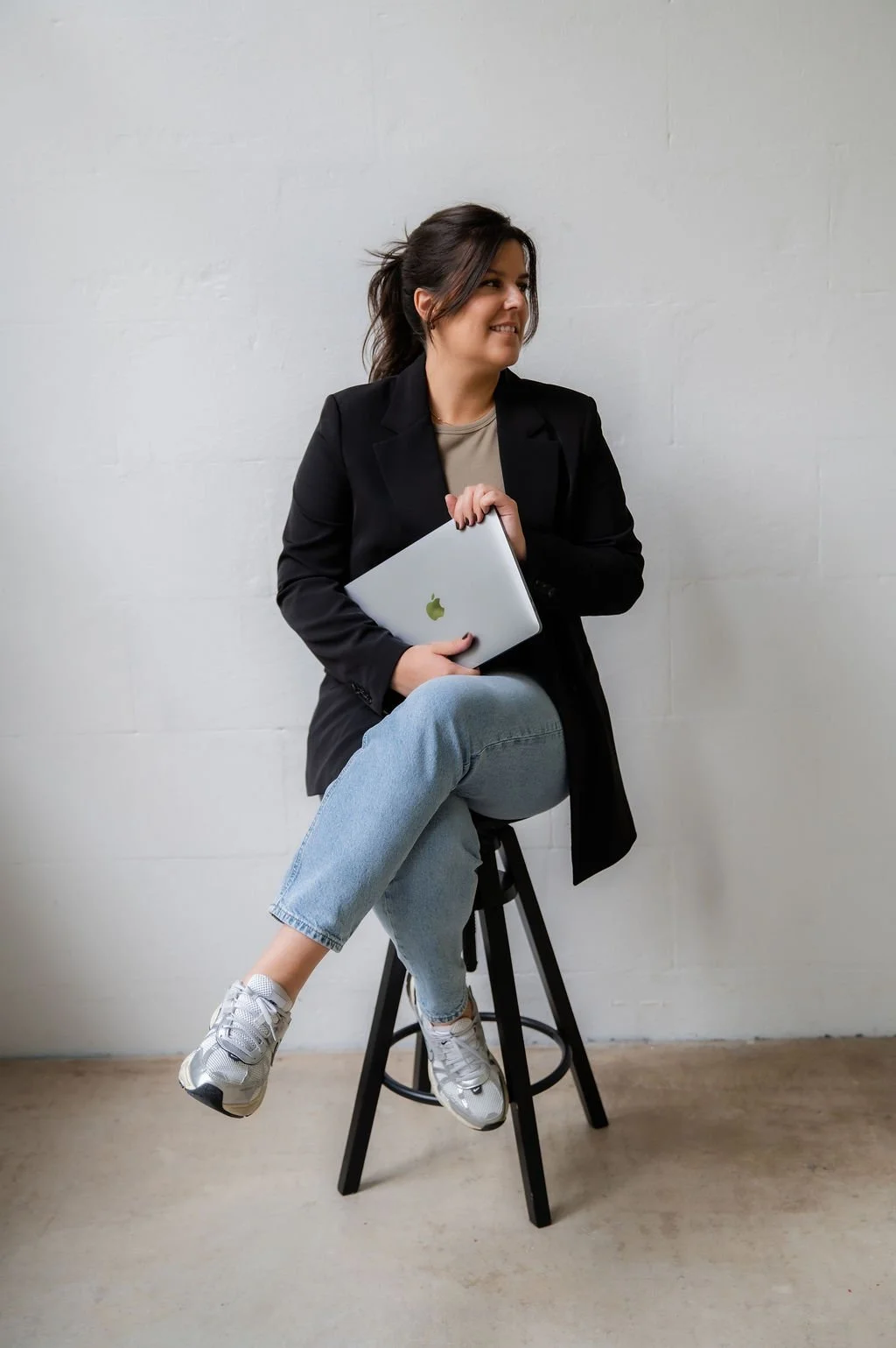 Woman sitting on a stool, holding a laptop, wearing a black blazer, jeans, and sneakers, against a plain wall.