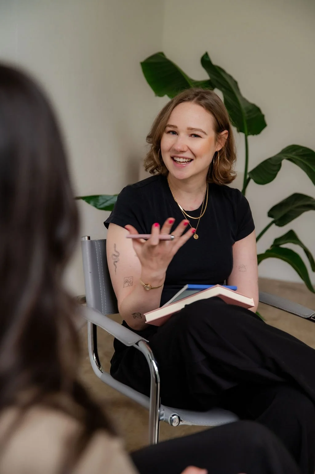 A woman with short brown hair smiling and talking, sitting in a chair with a notebook on her lap, wearing a black t-shirt, gold jewelry, and pink nail polish, with a large green plant in the background.