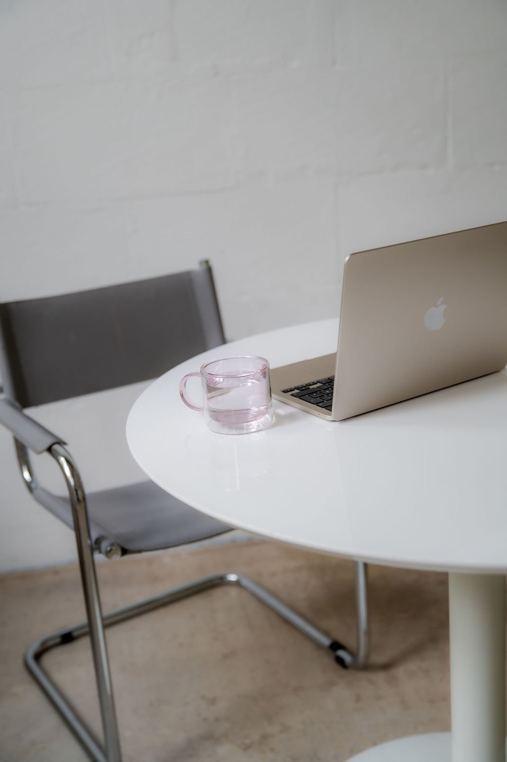 A round white table with a laptop and a transparent pink glass cup filled with water, accompanied by a modern chair with a gray cushion and metal armrests in a minimalistic setting.