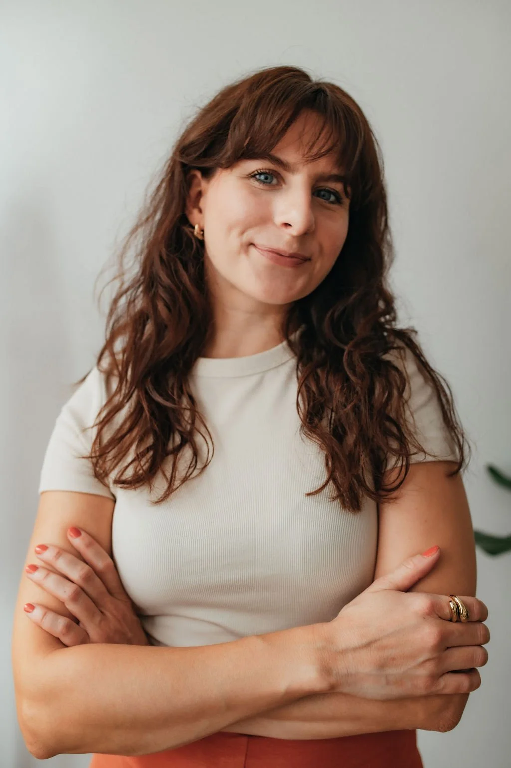 A woman with brown, curly hair and blue eyes stands with arms crossed, smiling at the camera, wearing a white short-sleeved shirt and gold jewelry, in front of a plain light-colored background.