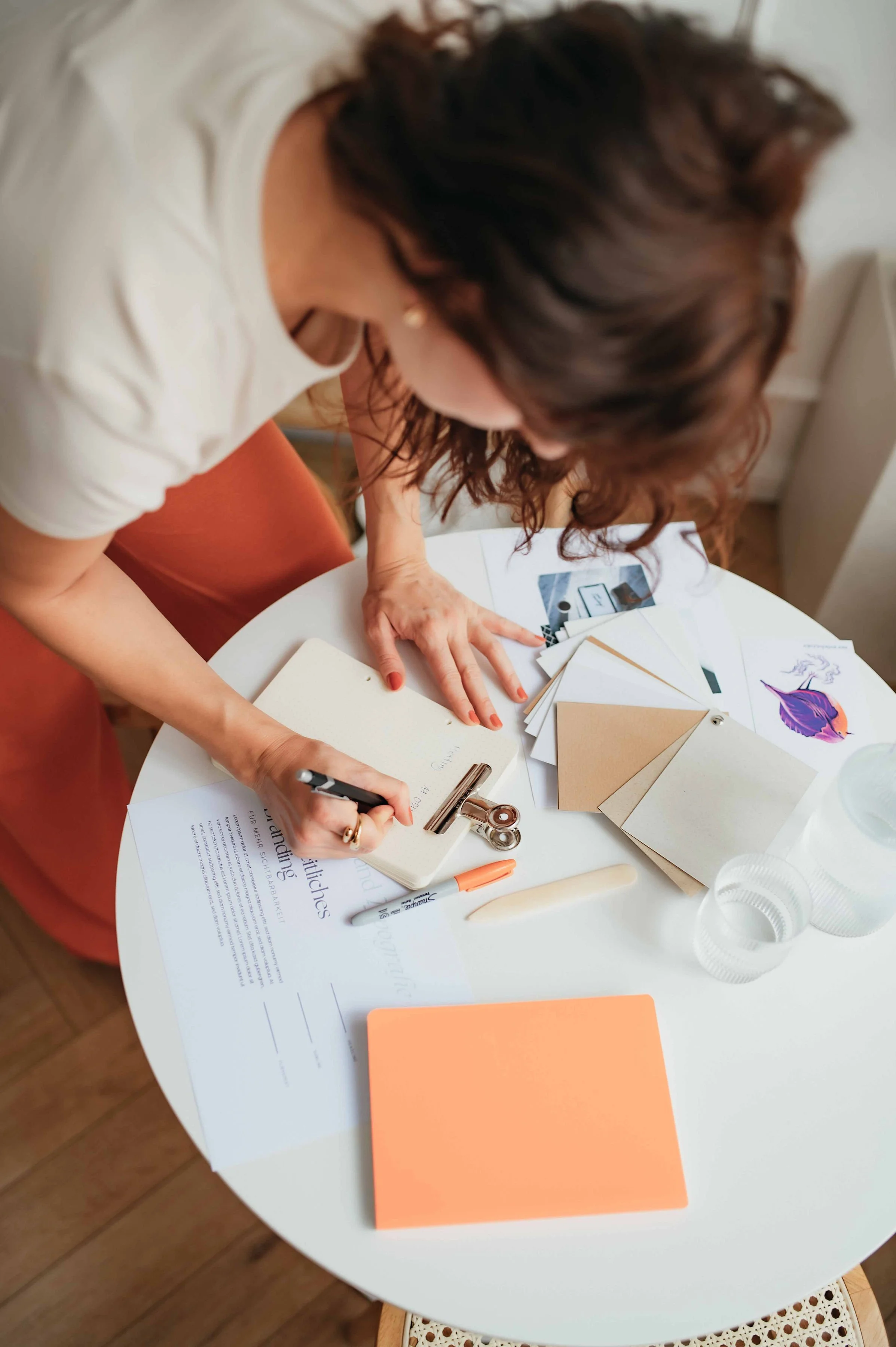 A woman working at a small round white table with various papers, colored cards, and office supplies scattered across it.