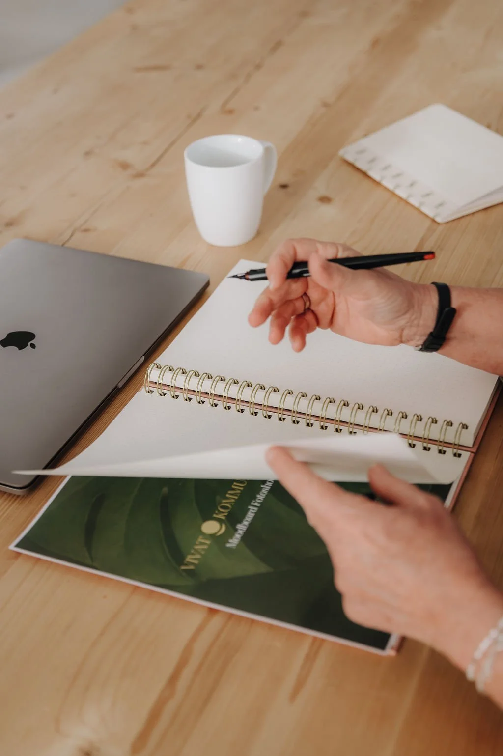 A person writing in a spiral notebook at a wooden table with a MacBook, a white mug, a stack of napkins, and a folder.