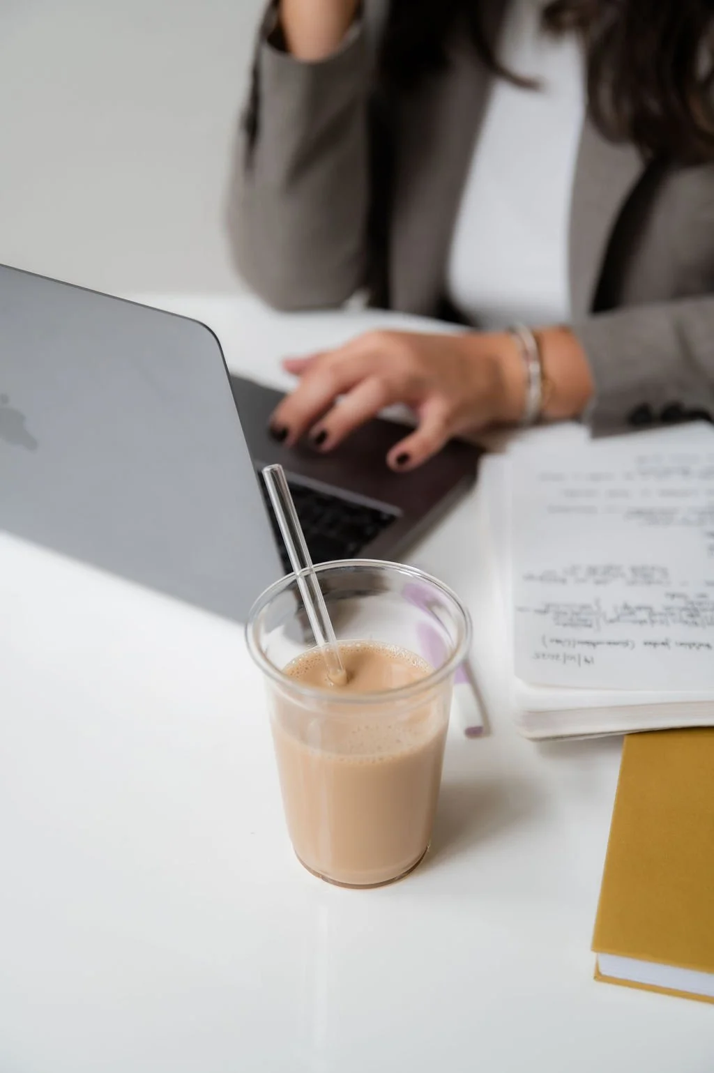A glass of milk tea with a straw on a white table, with a woman in a gray blazer working on a laptop and an open notebook nearby.