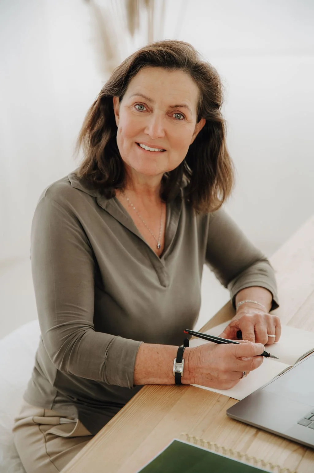 A woman with shoulder-length brown hair, smiling, sitting at a wooden desk with a laptop, notebook, and pen, in a bright room.