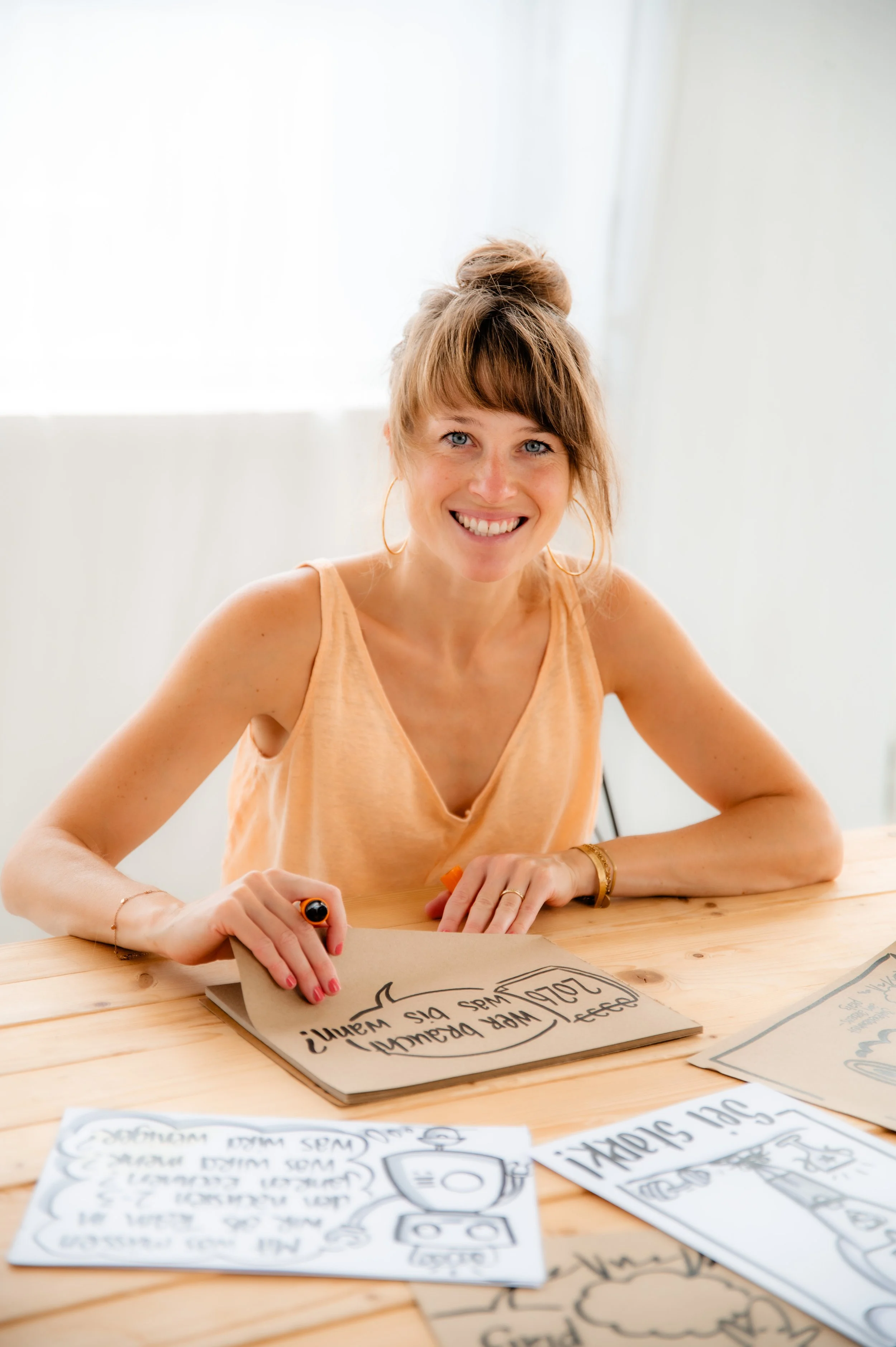 A woman with light brown hair tied up, wearing a sleeveless peach top, sitting at a wooden table and smiling while drawing comic-style sketches on papers.