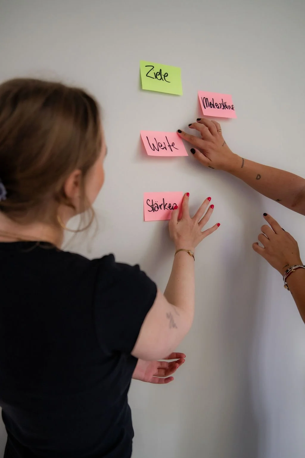 Two women are placing colorful sticky notes on a white wall. The notes have German words written on them, including 'Ziel,' 'Medikamente,' 'Welt,' and 'Stärke.'