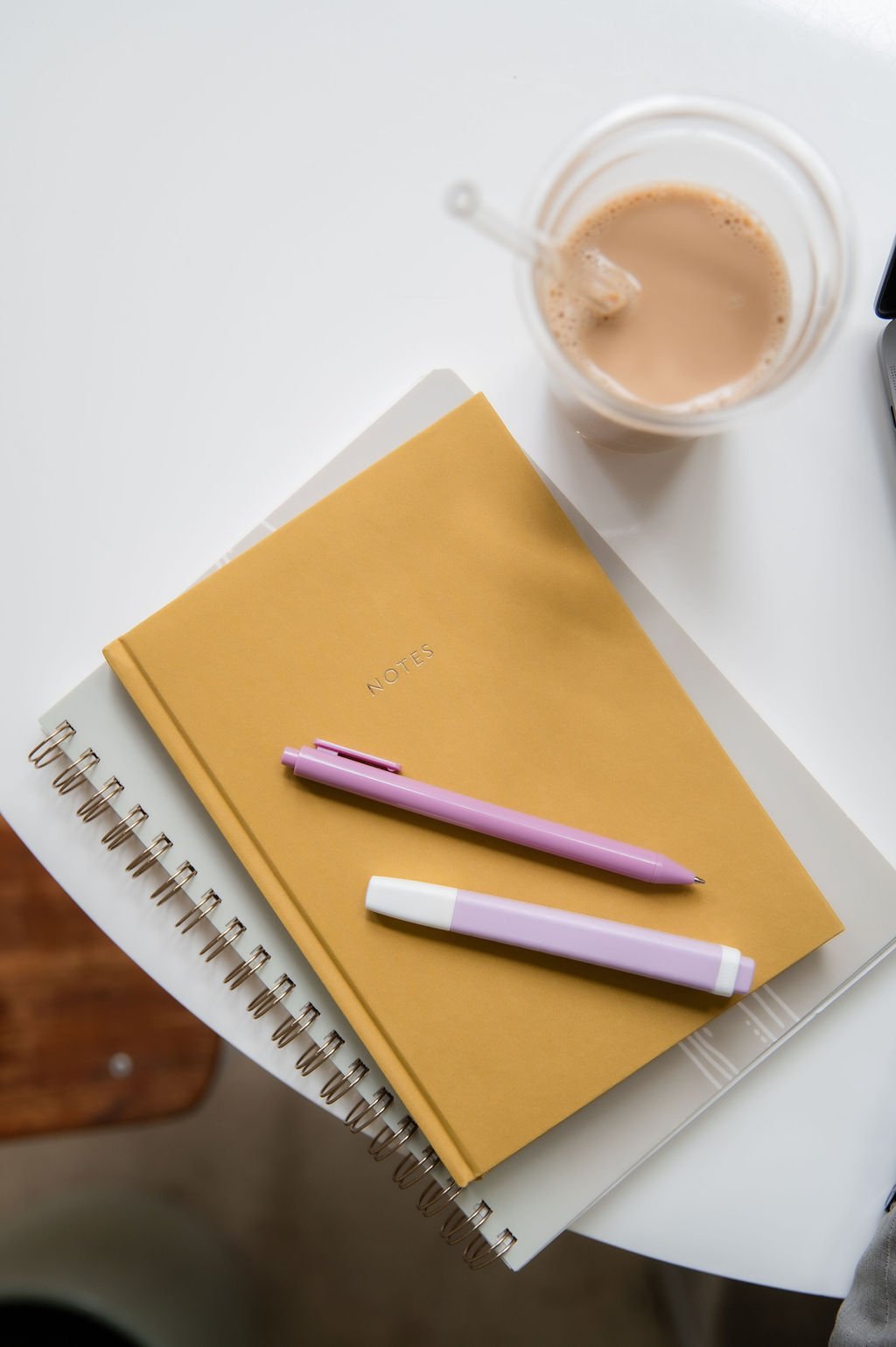 A white round table holding a yellow notebook, a pink pen, a white and pink highlighter, a clear glass of iced coffee with a straw, and part of a laptop.
