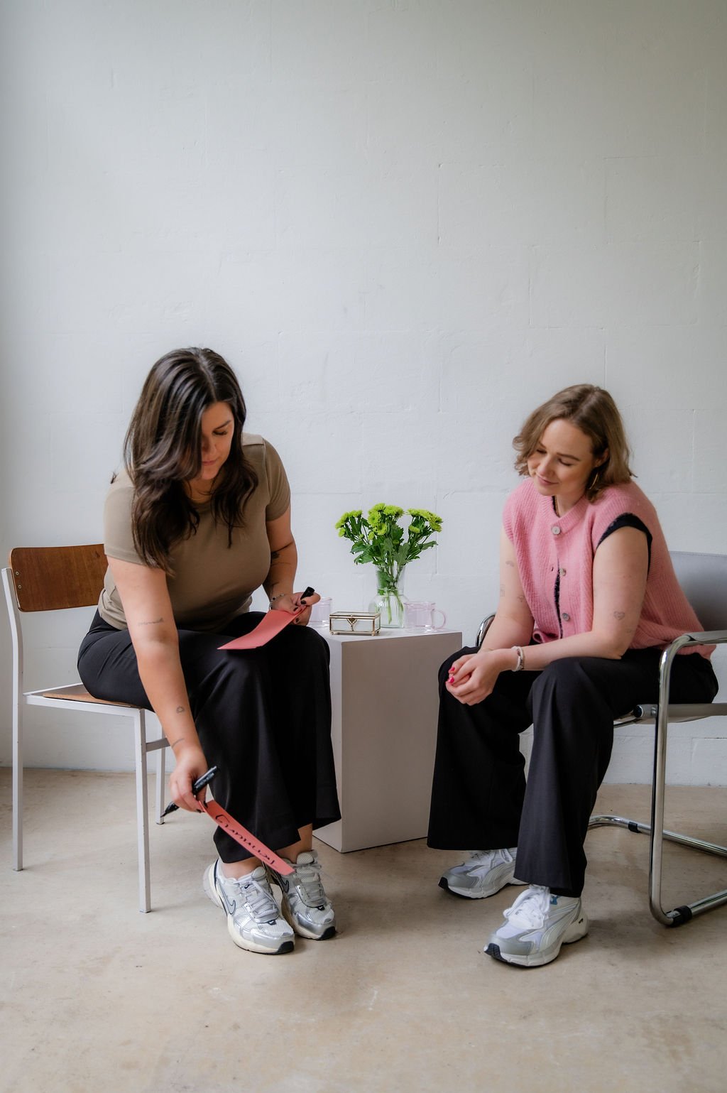 Two women sitting in a room with a white brick wall. One woman is holding a pink paper, and the other woman is sitting across from her, smiling. A small table with a vase of green flowers is between them.