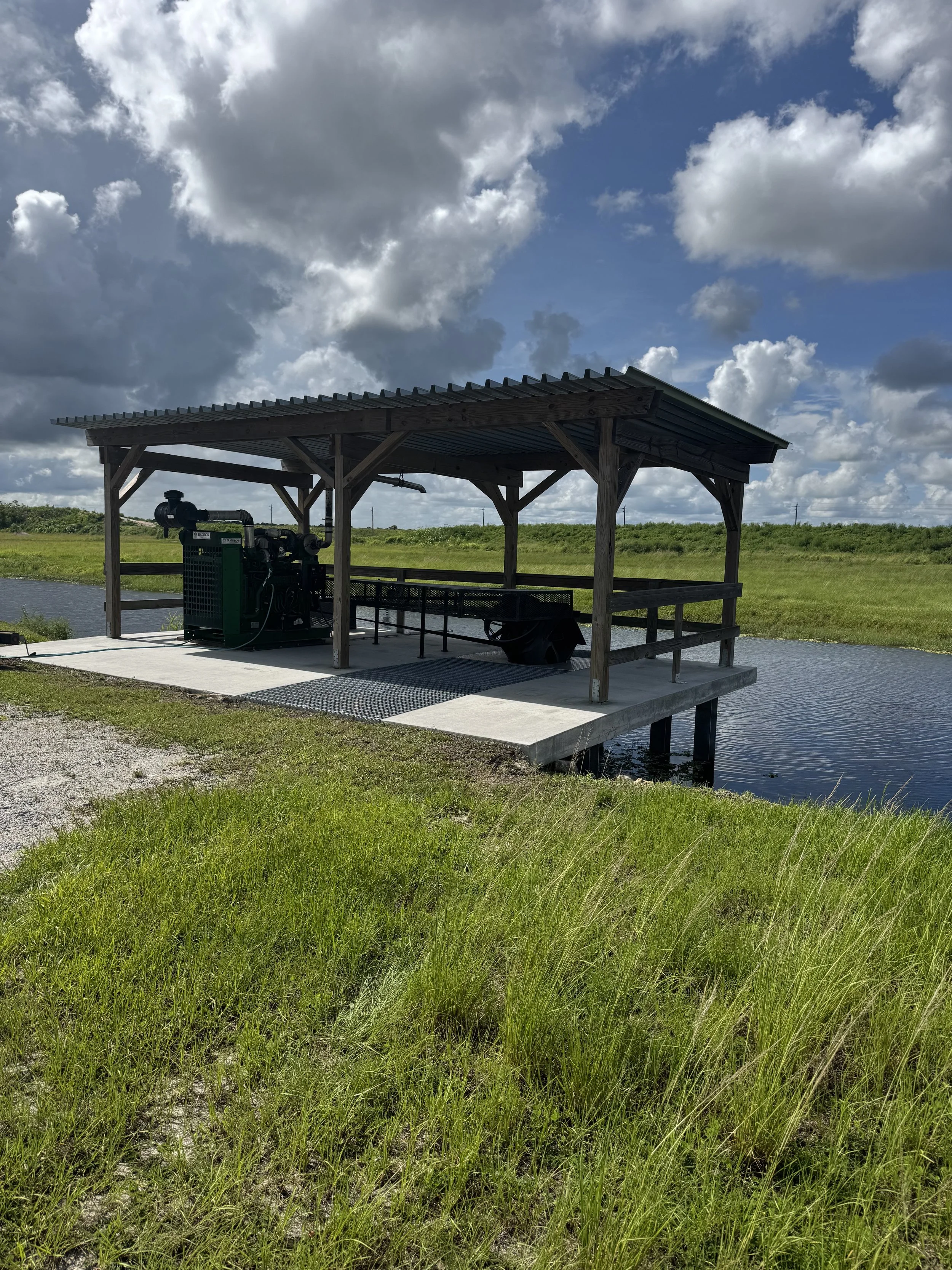 A small covered structure with a generator and picnic table, situated on a concrete platform over a body of water with grassy surroundings and a partly cloudy sky.