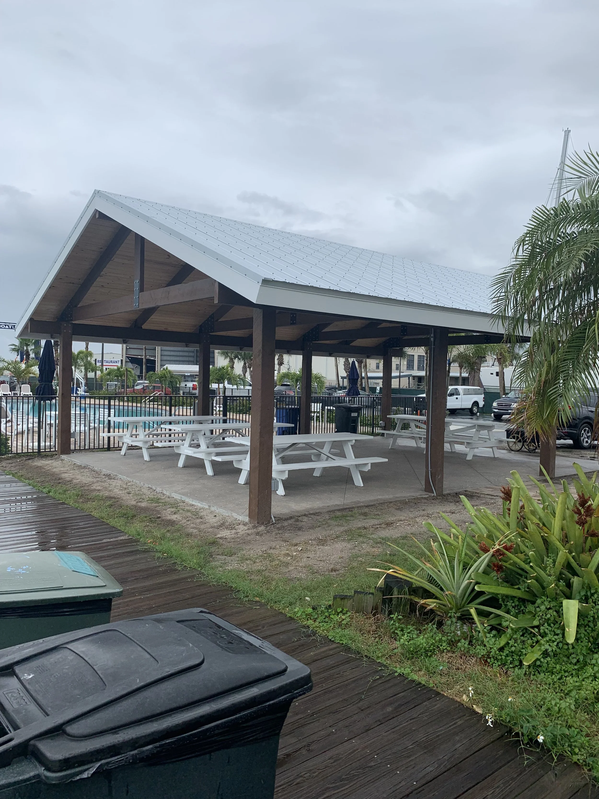 Outdoor pavilion with a metal roof and white picnic tables, surrounded by plants and nearby parking lot with cars, under cloudy sky.