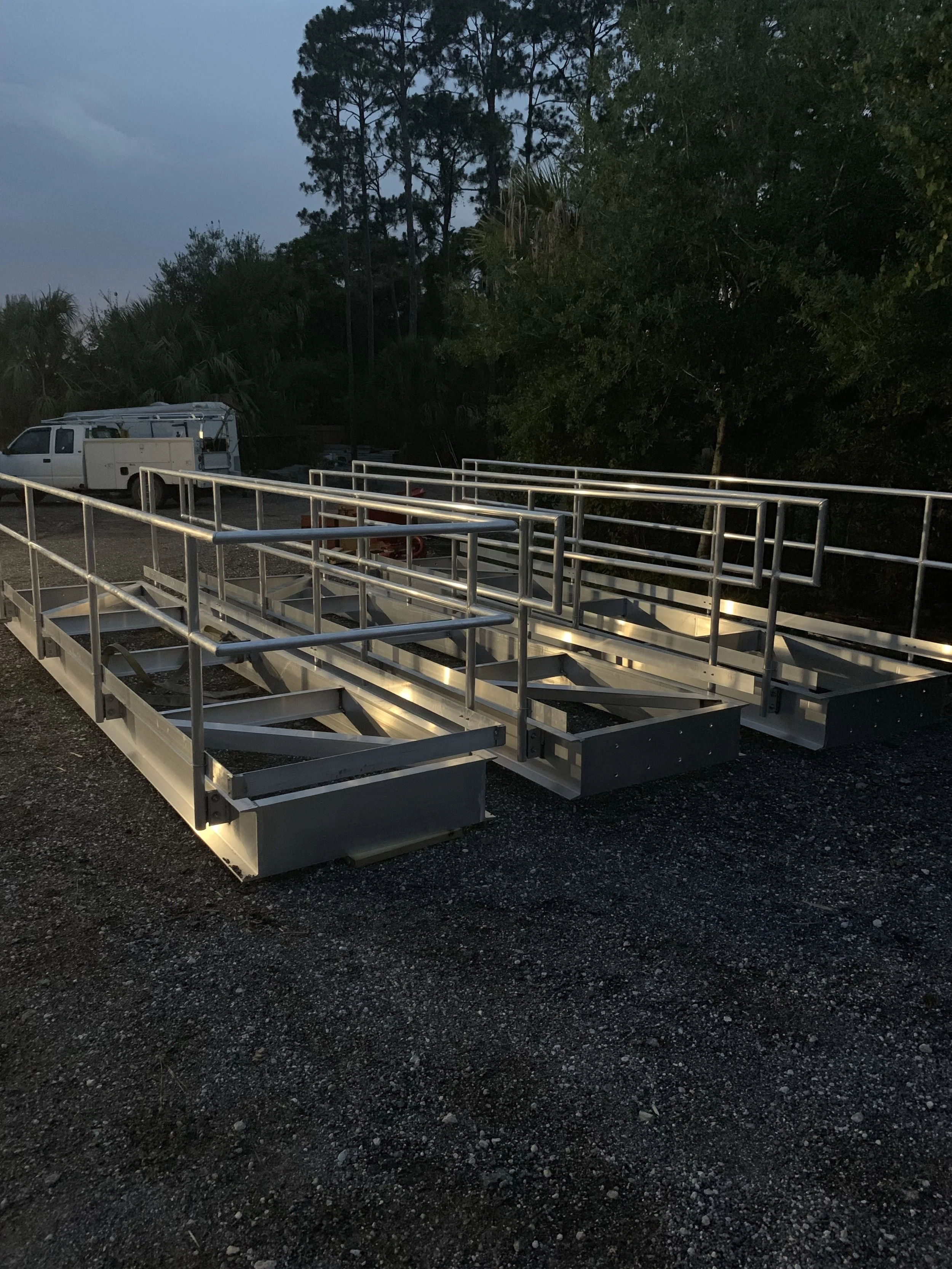 Stainless steel mobile staircase with handrails, positioned outdoors on gravel, with trees and a white utility truck in the background, during evening or early morning.