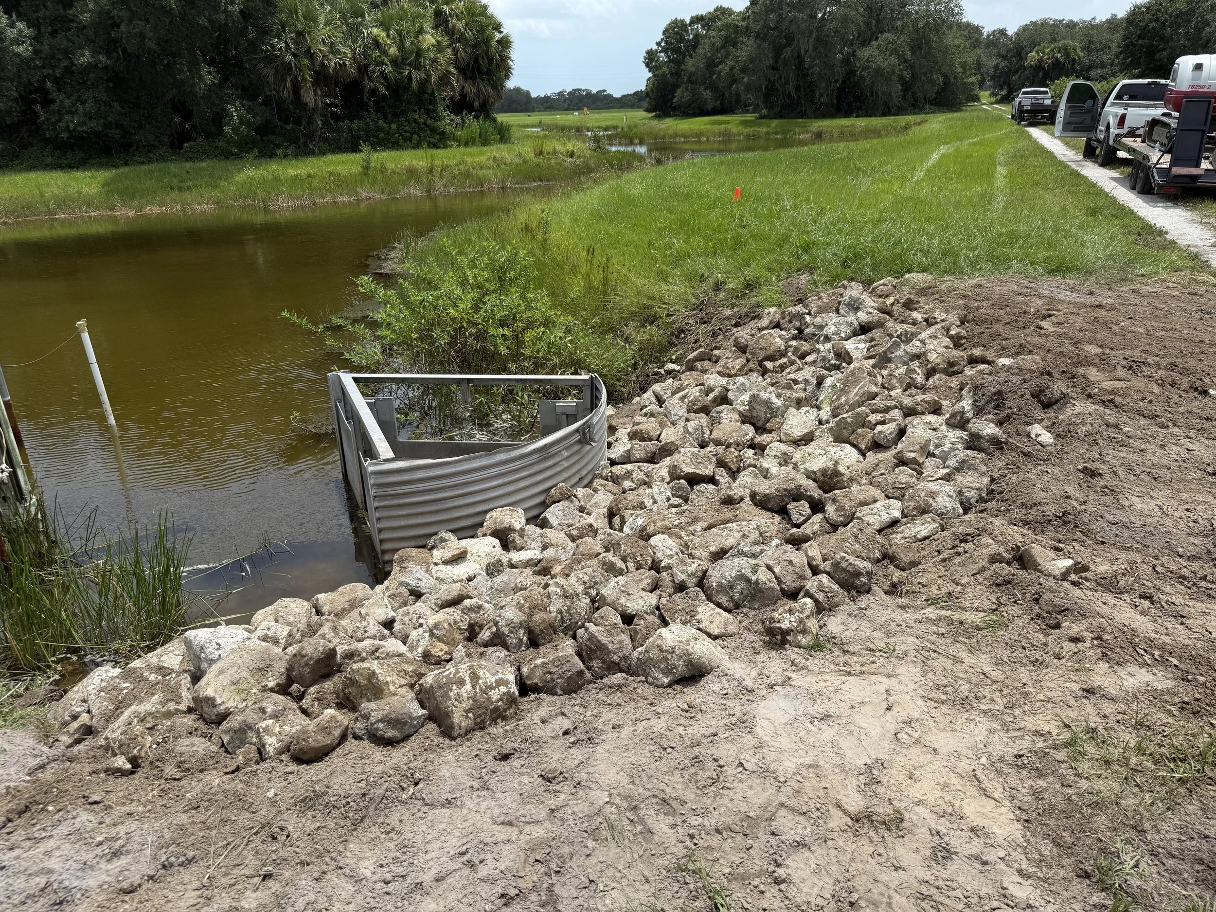 New rocks placed along the edge of a waterway with a metal barrier at the corner, and several parked trucks and vehicles on a dirt road beside a grassy field, under a mostly cloudy sky.