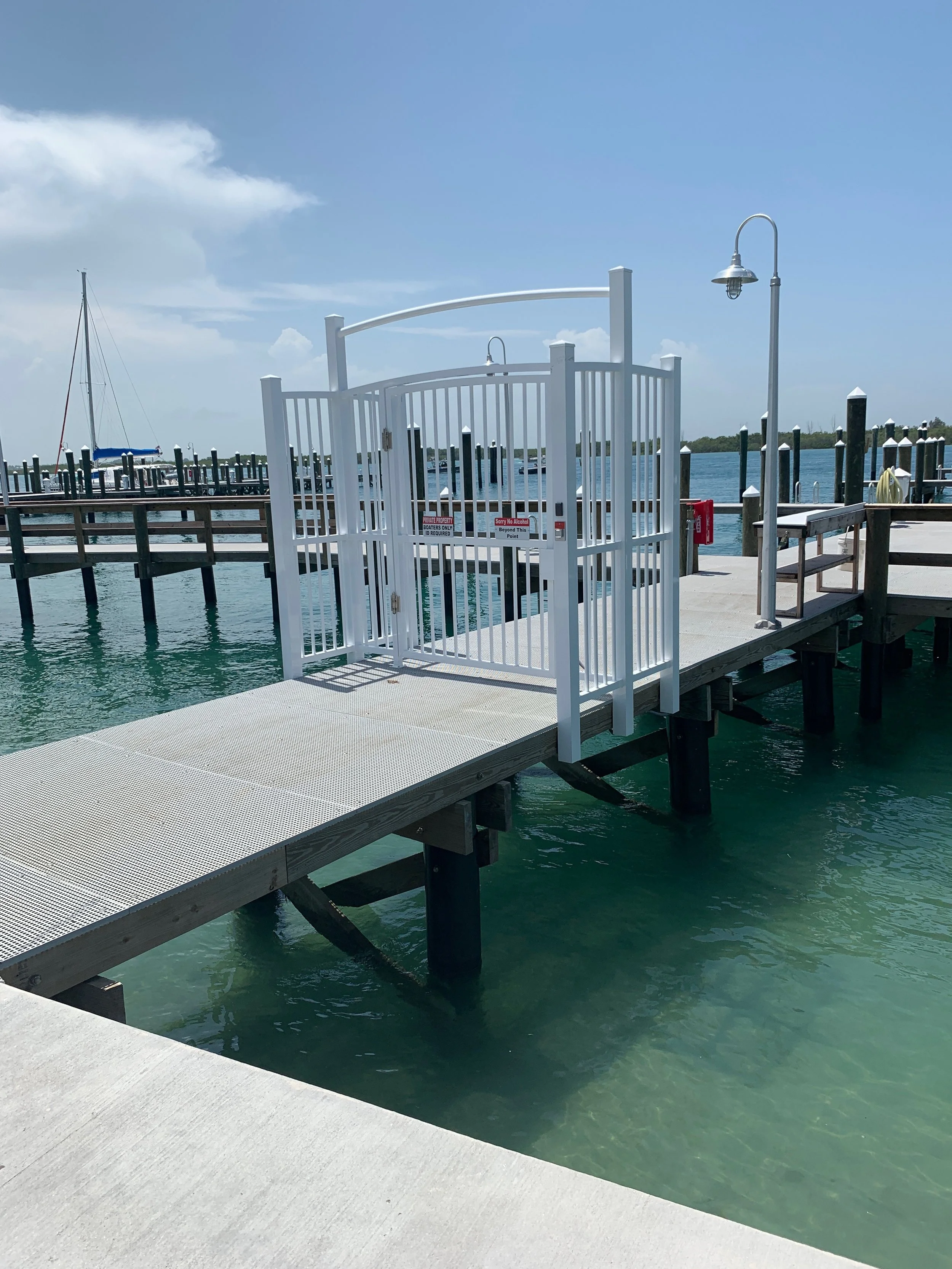 A marina with a gate on the dock, boats, and a lamppost under a partly cloudy sky.
