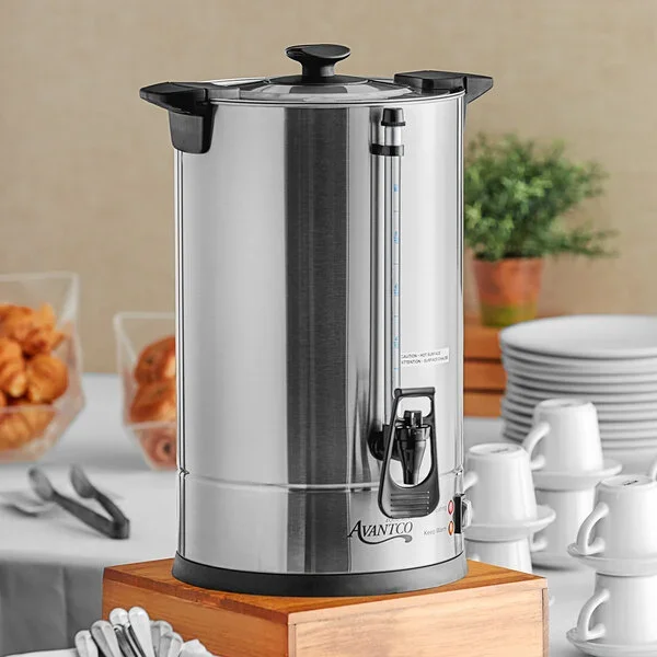 A stainless steel water boiler on a wooden tray, with a water level indicator, spout, and black lid, surrounded by plates and cups in a buffet setting.