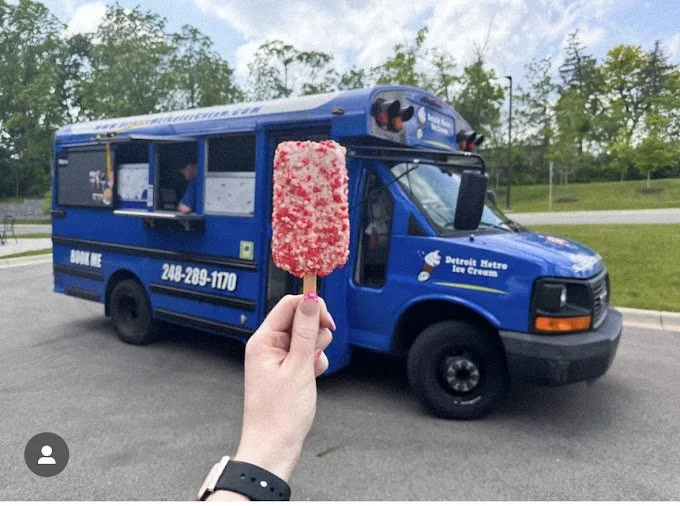 Person holding a pink and white ice cream bar in front of a blue Detroit Metro Ice Cream truck parked on a road.